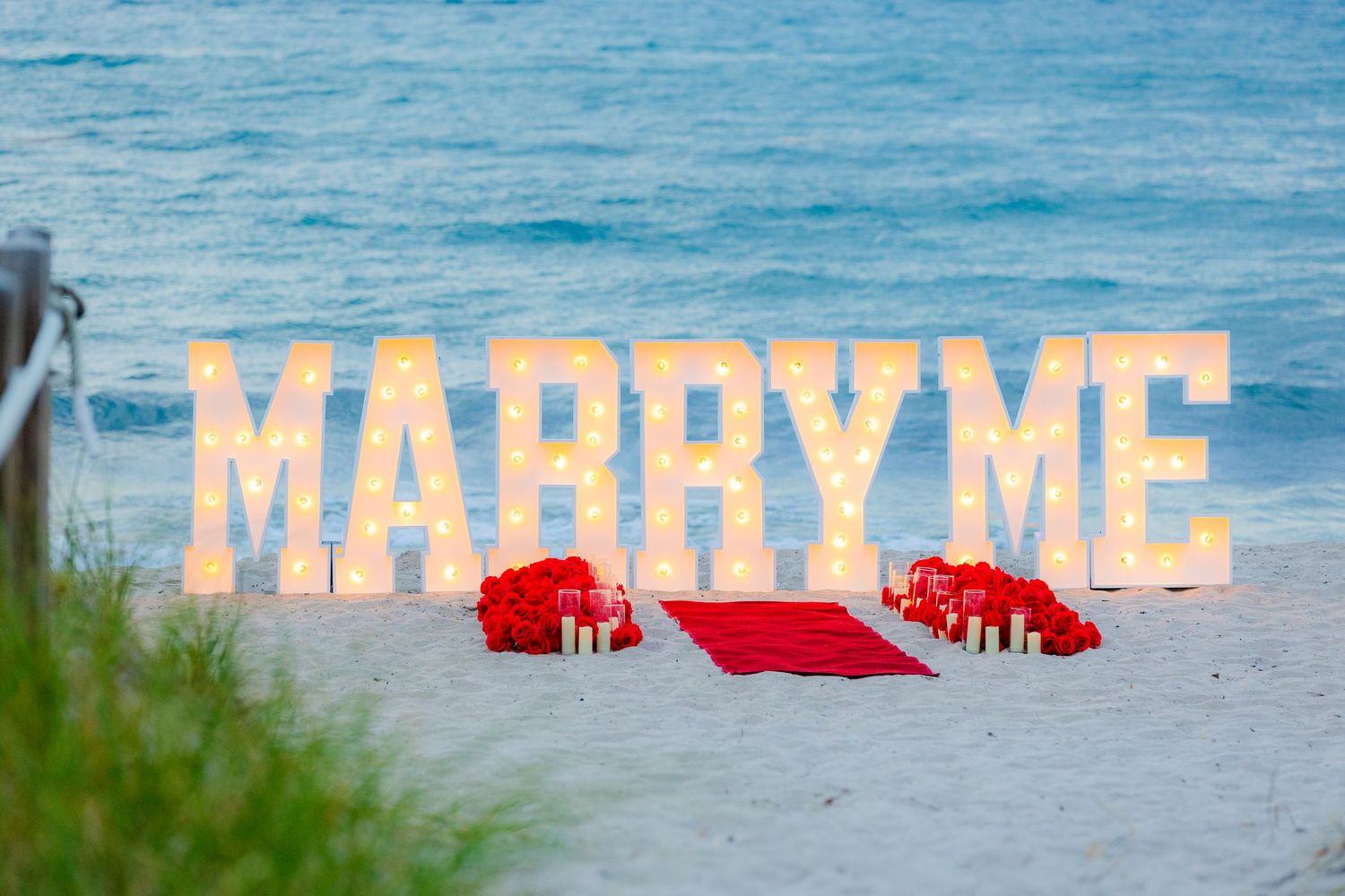 Illuminated letters spelling out 'MARRY ME' set up on a sandy beach with blue ocean water in the background.