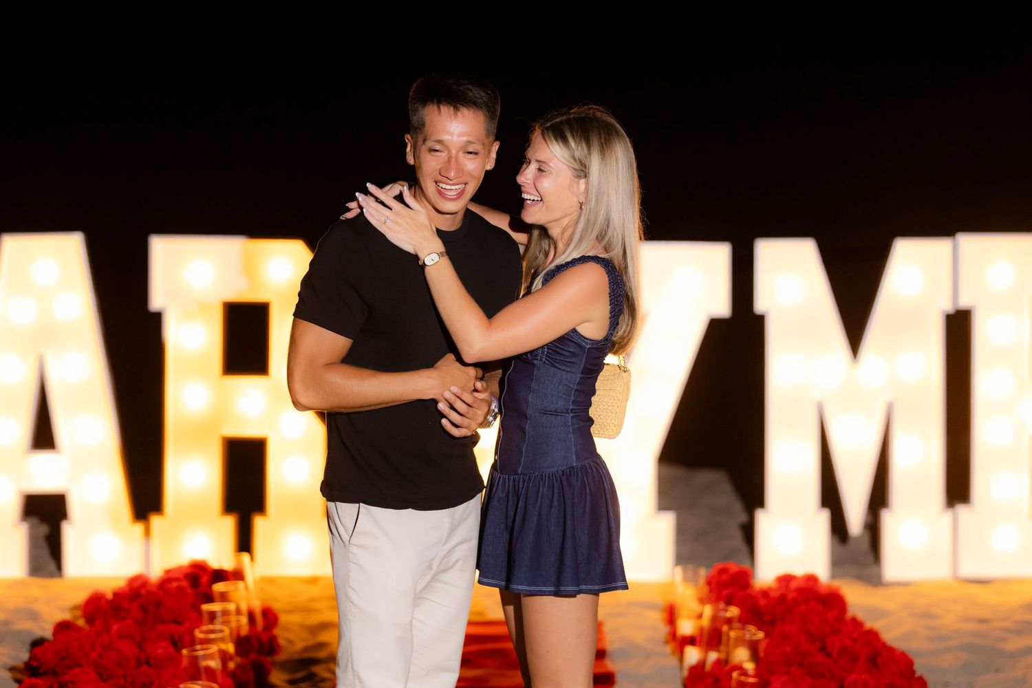 Couple sharing a tender moment during nighttime proposal with glowing 'MARRY ME' sign in background.