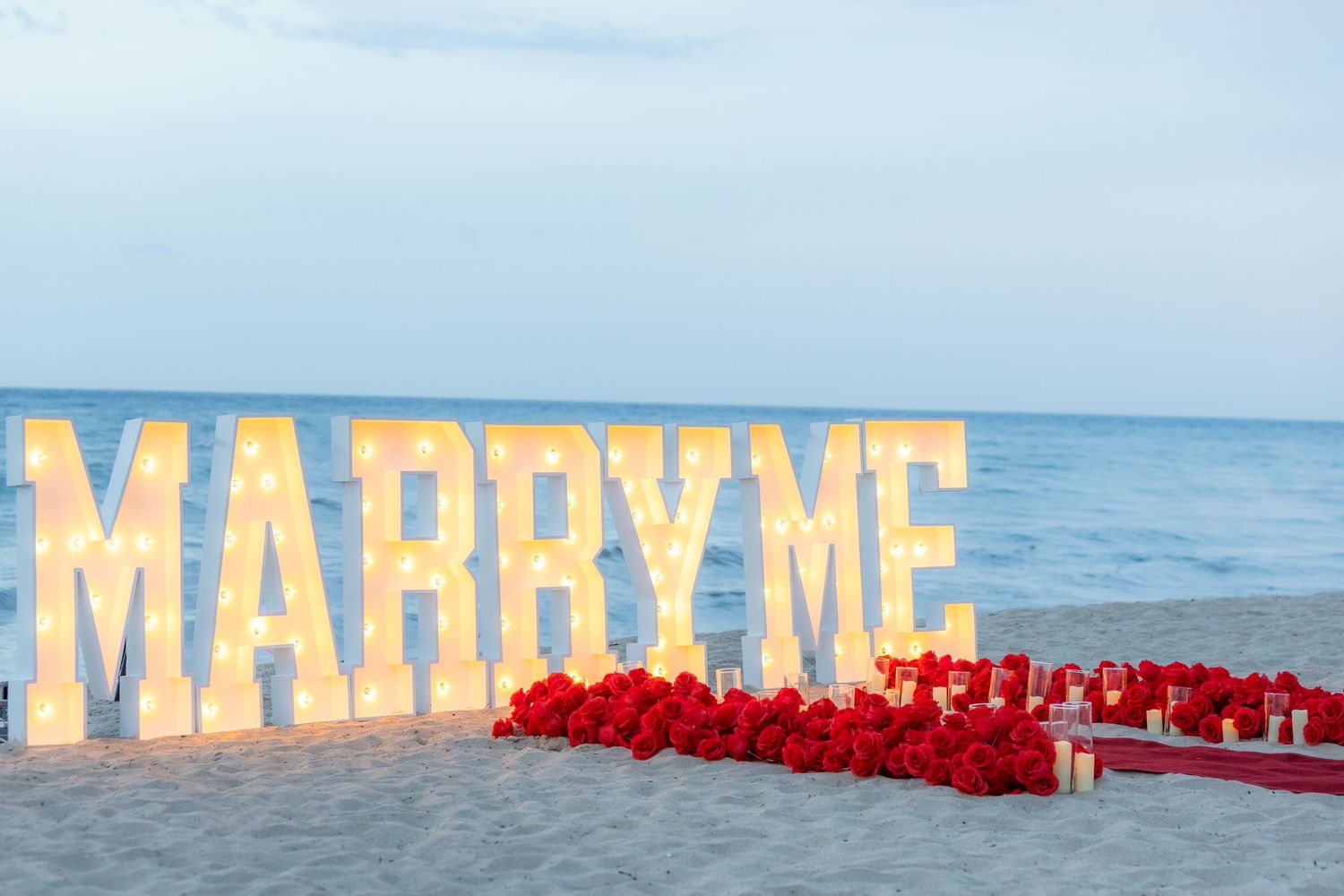 Large illuminated "MARRY ME" letters display a romantic beach proposal setup with red roses and ocean backdrop.
