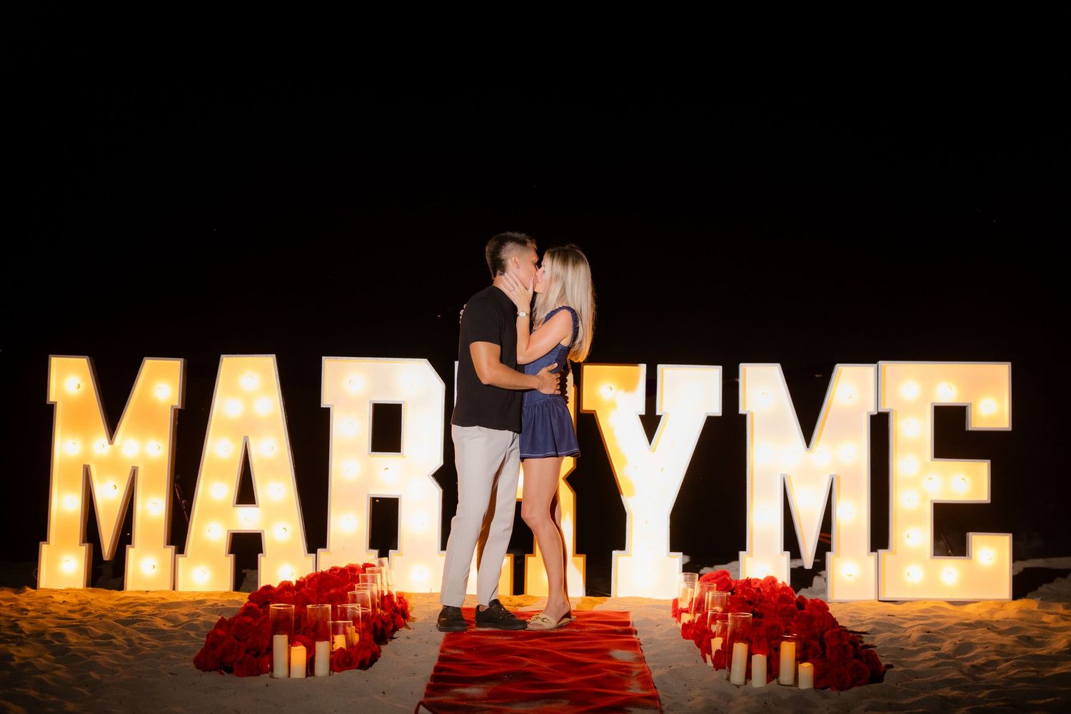 Silhouetted couple embracing near glowing MARRY ME letters surrounded by red rose petals at night.