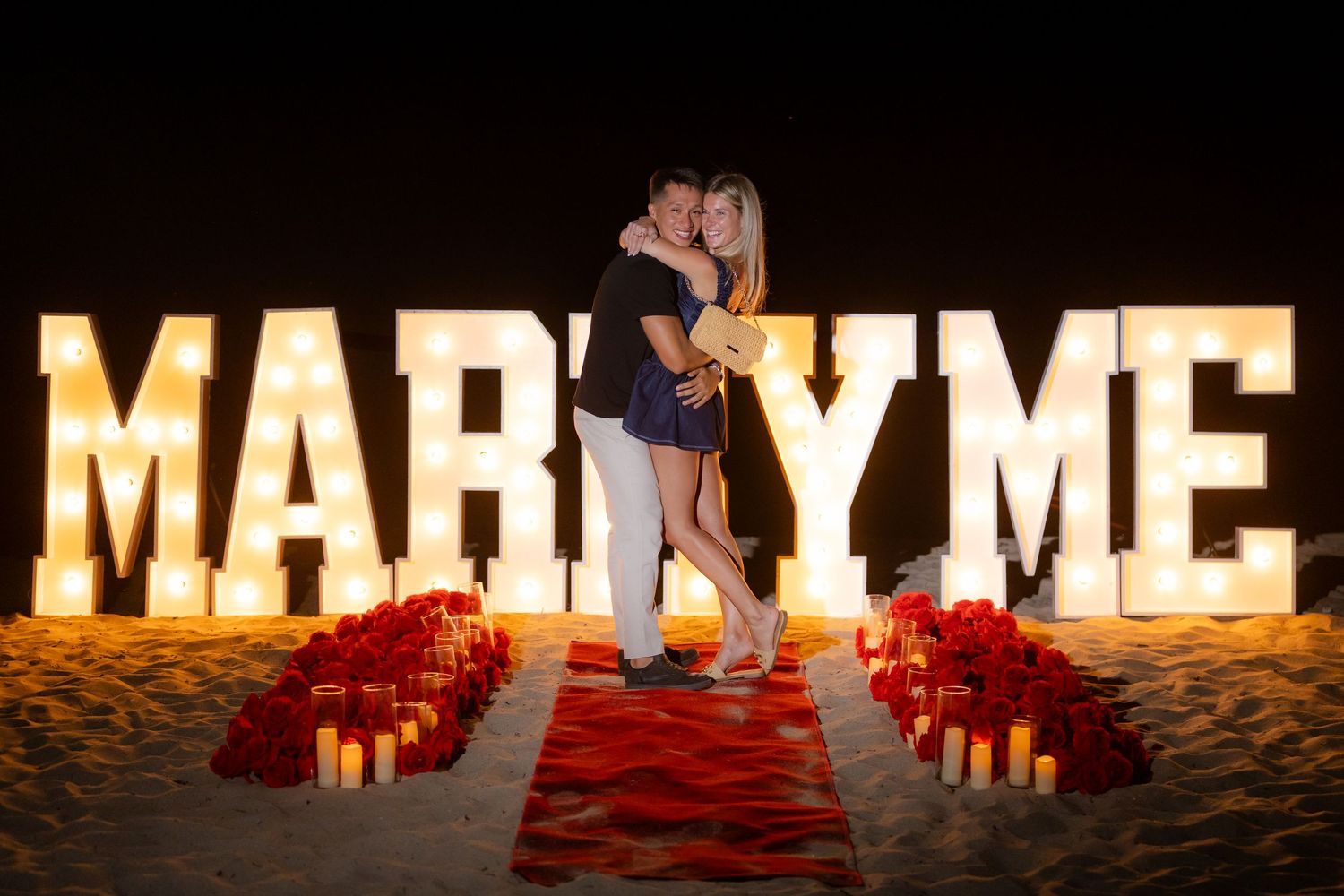 Illuminated letters spelling 'MARRY ME' glow warmly against a dark background in a romantic proposal setup.