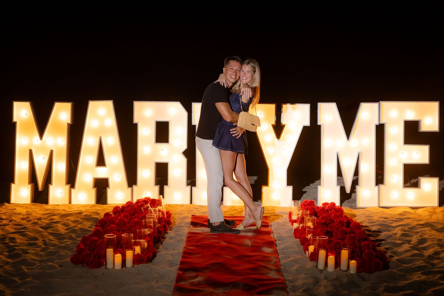 Illuminated letters spell out 'MARRY ME' at sunset with candles set up for romantic beach proposal.