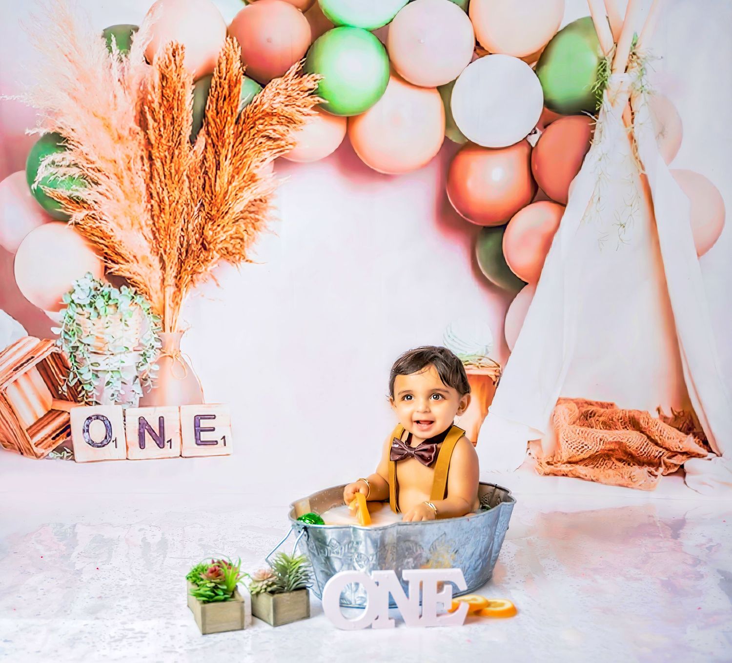 A whimsical party setup with coral and mint colored balloons, dried pampas grass, and wooden letter decorations against white.