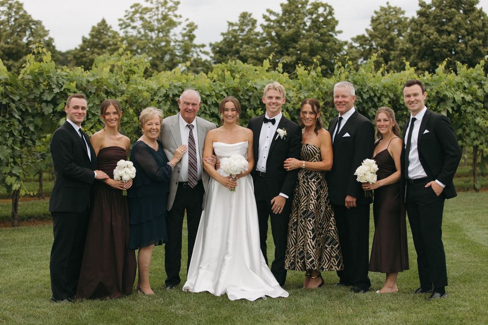 Elegant wedding party poses together on lawn with trees and greenery in background.