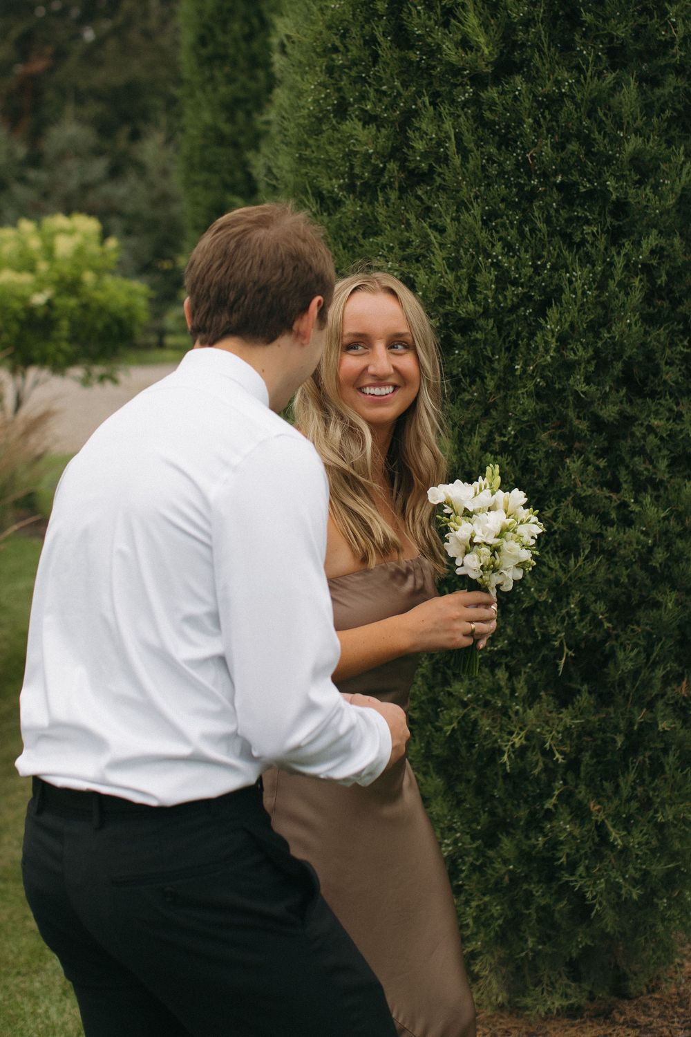Candid moment between couple sharing flowers by garden hedge.