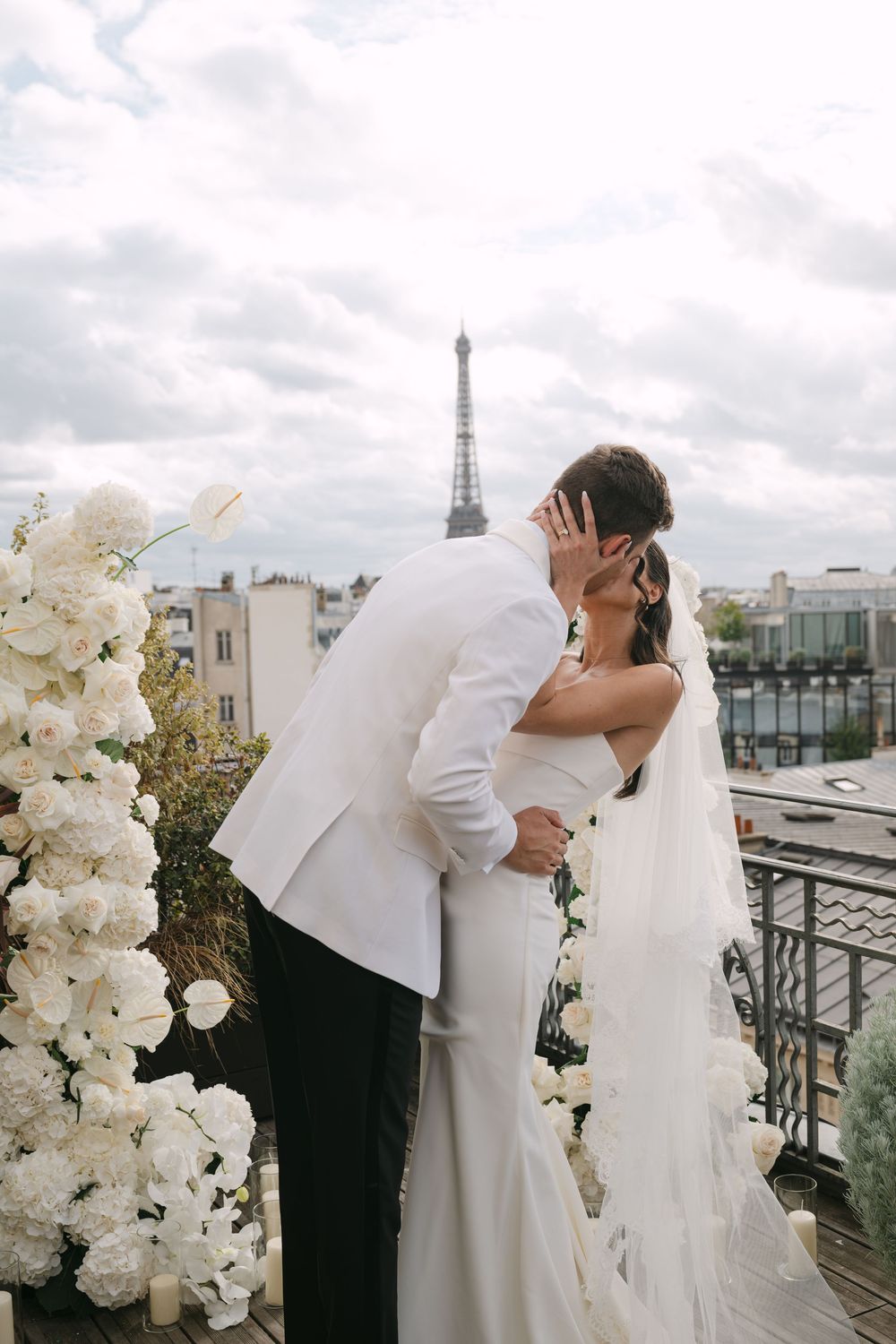 A romantic wedding couple embraces on a Parisian rooftop terrace with white floral decorations and the Eiffel Tower visible.