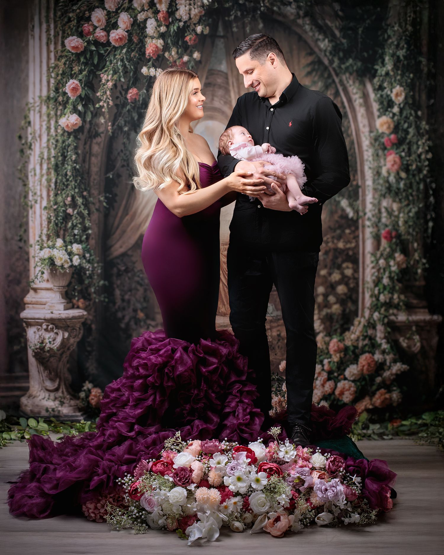 A family portrait the mother wearing a deep purple designer gown the father wearing black holding their daughter wearing a mauve tulle dress