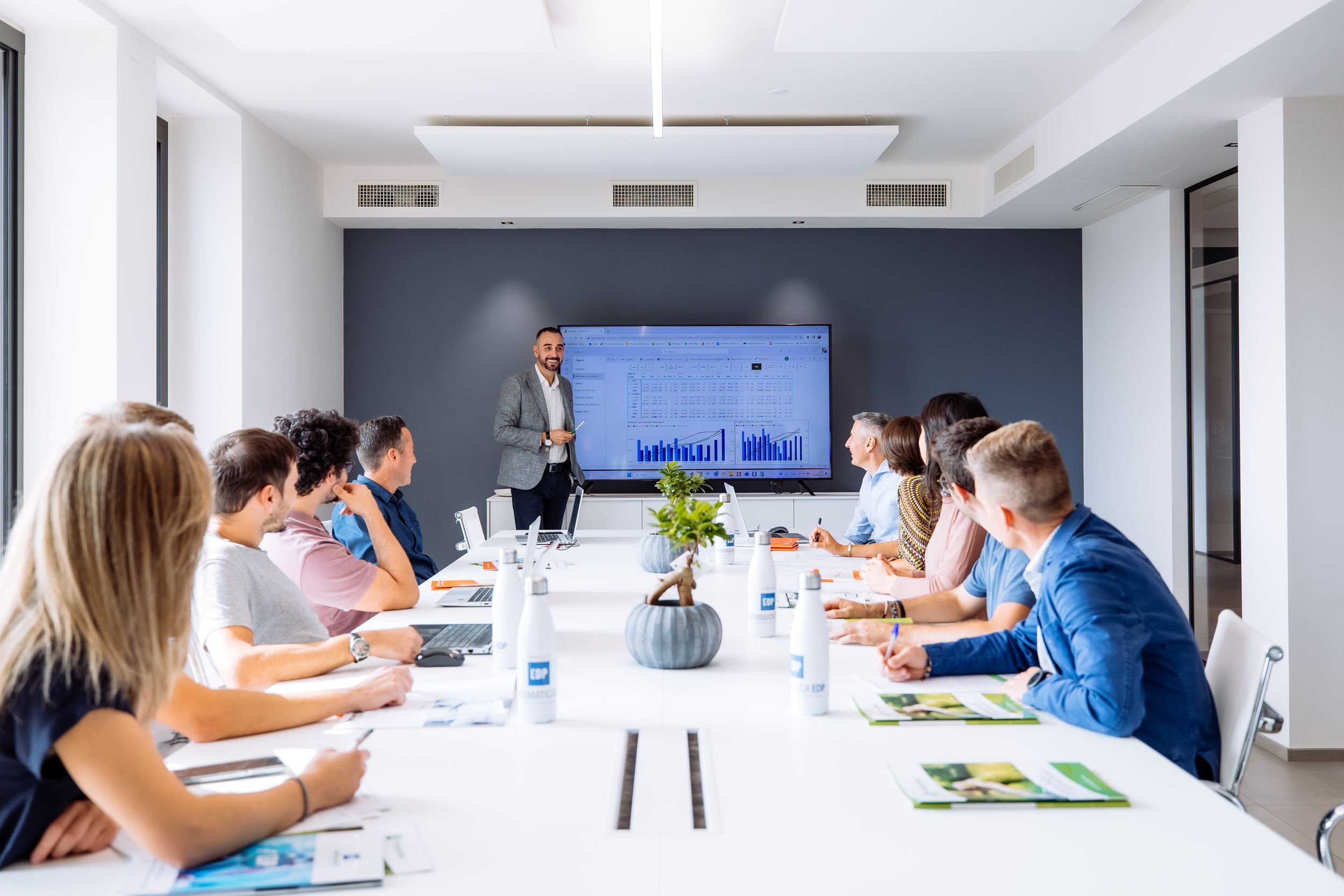 Business meeting in modern conference room with people seated around white table and presenter standing by blue wall.
