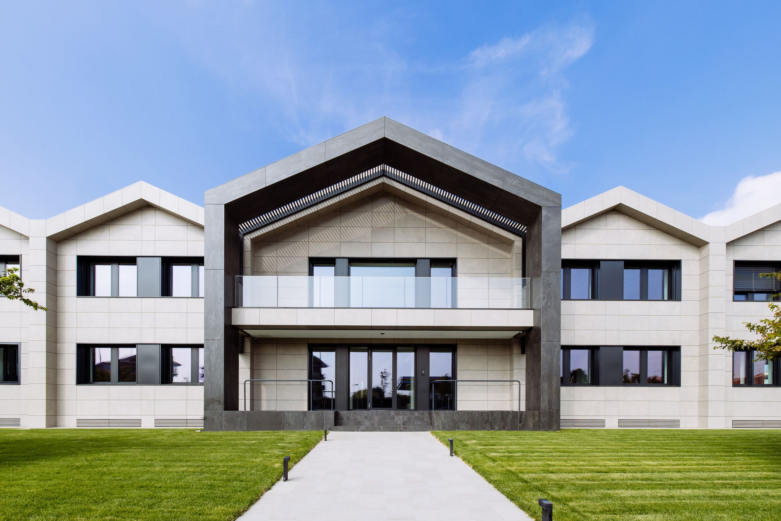 Modern contemporary rural houses with large glass windows and peaked roofs form a clean row against green grass and blue sky.
