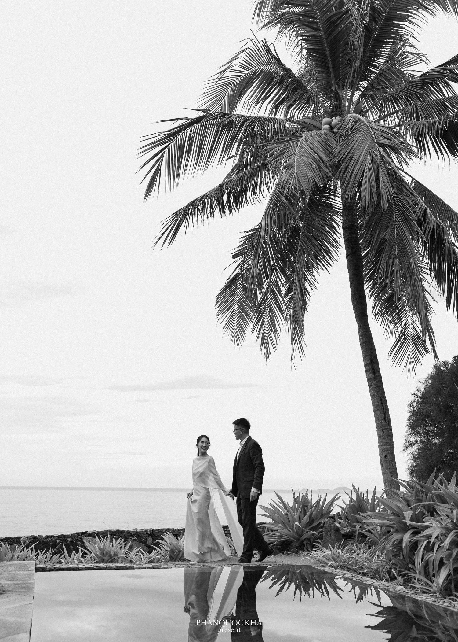 Black and white photo of couple standing by a pool under a palm tree near the ocean.