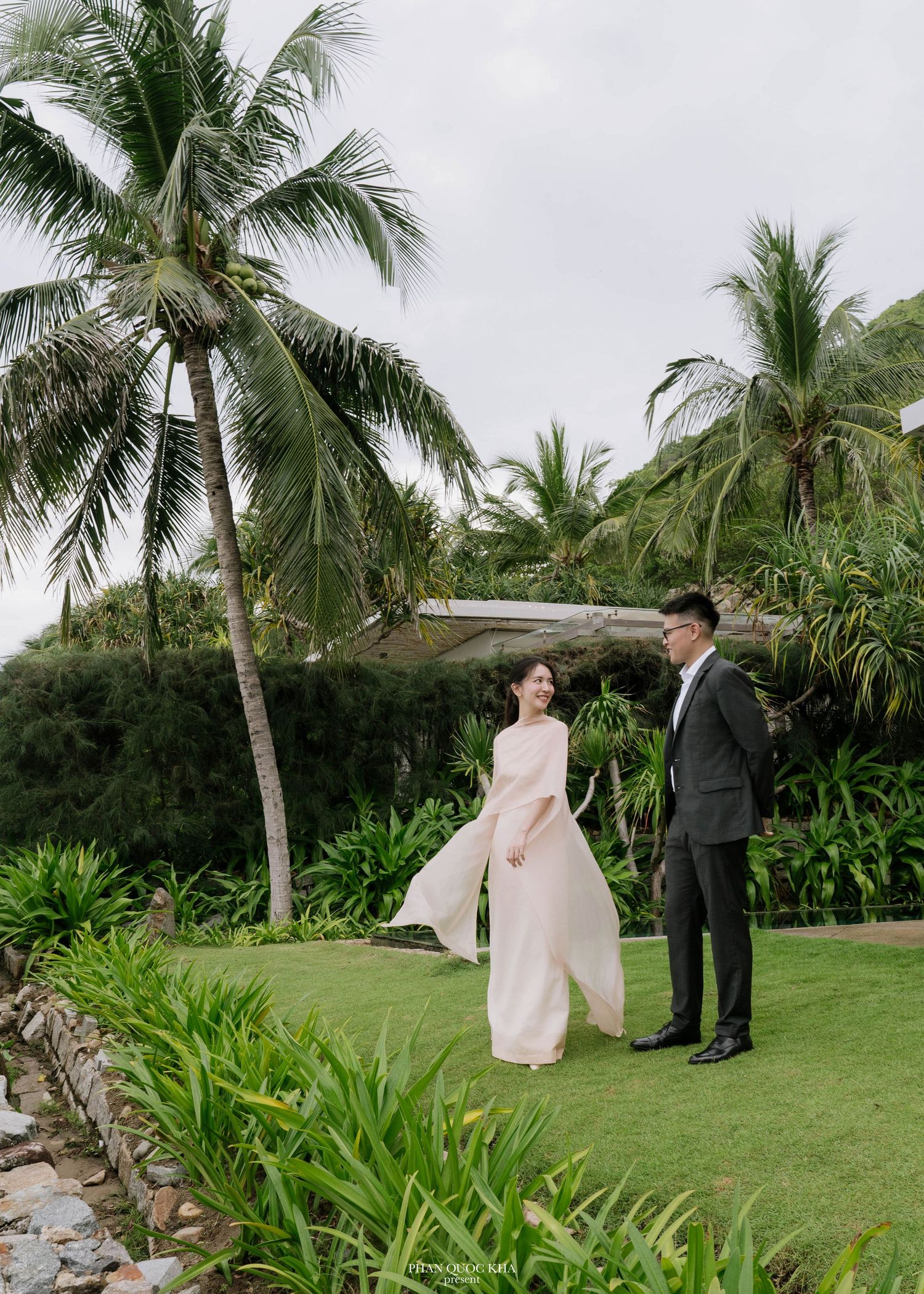 Bride in flowing pink gown and groom in dark suit on a tropical garden lawn.