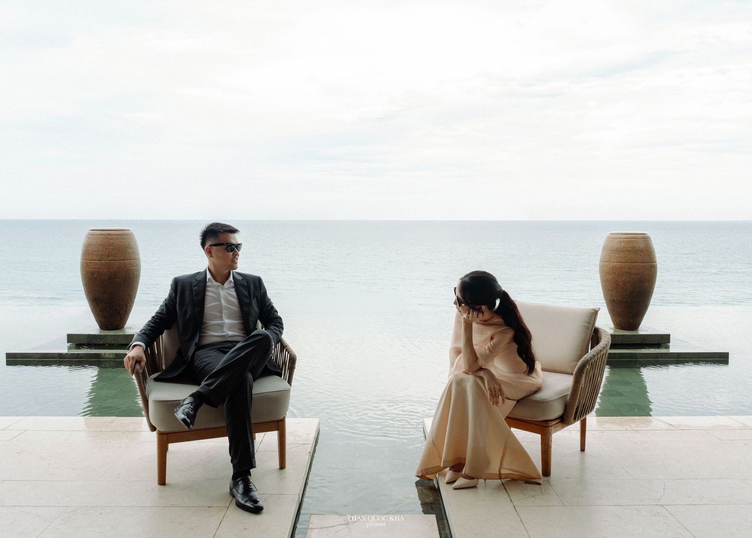 Couple sitting apart in elegant chairs by infinity pool with ocean view during pre-wedding shoot.