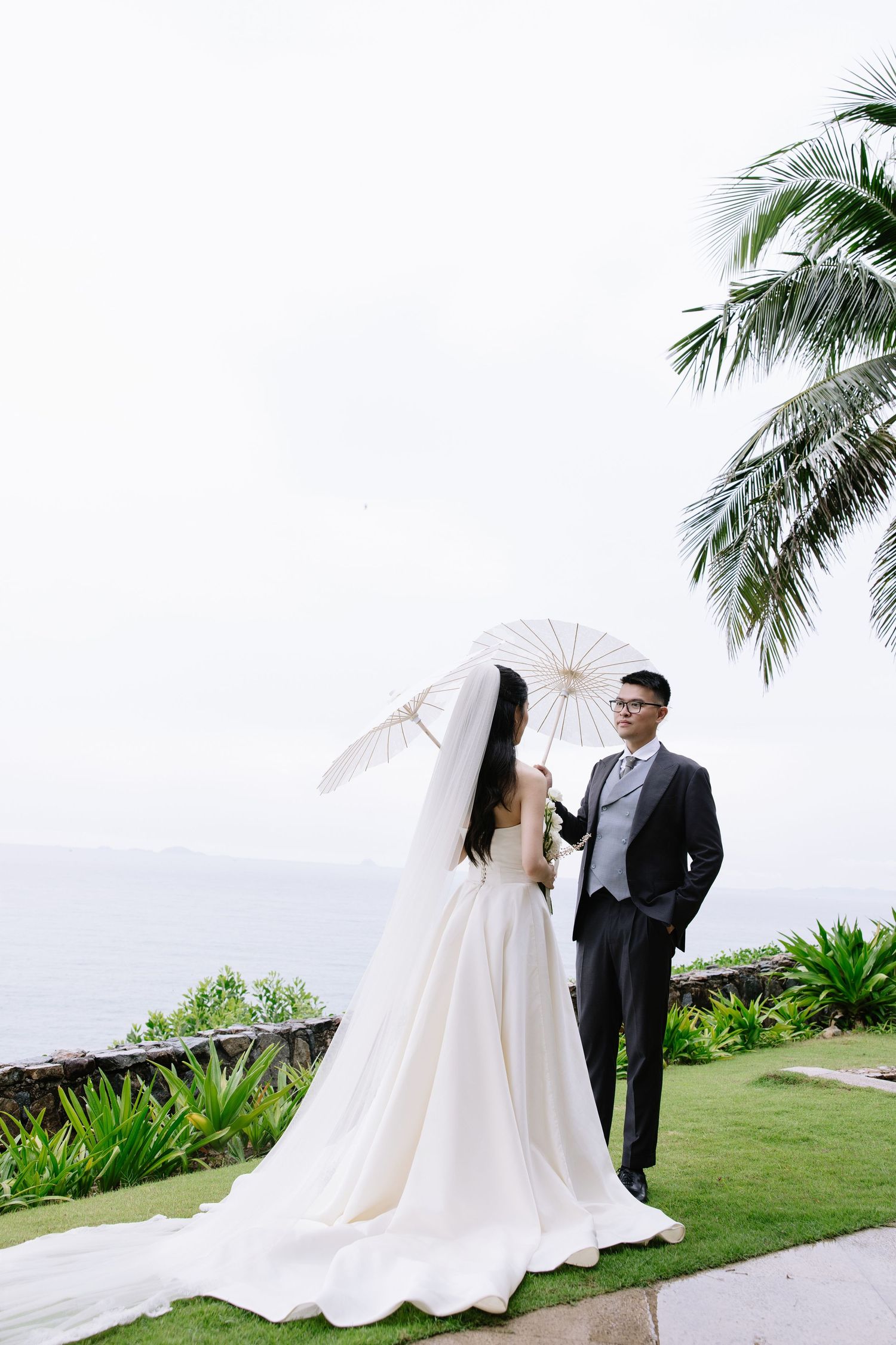 Bride holding a parasol and groom in dark suit standing on grass with ocean backdrop.