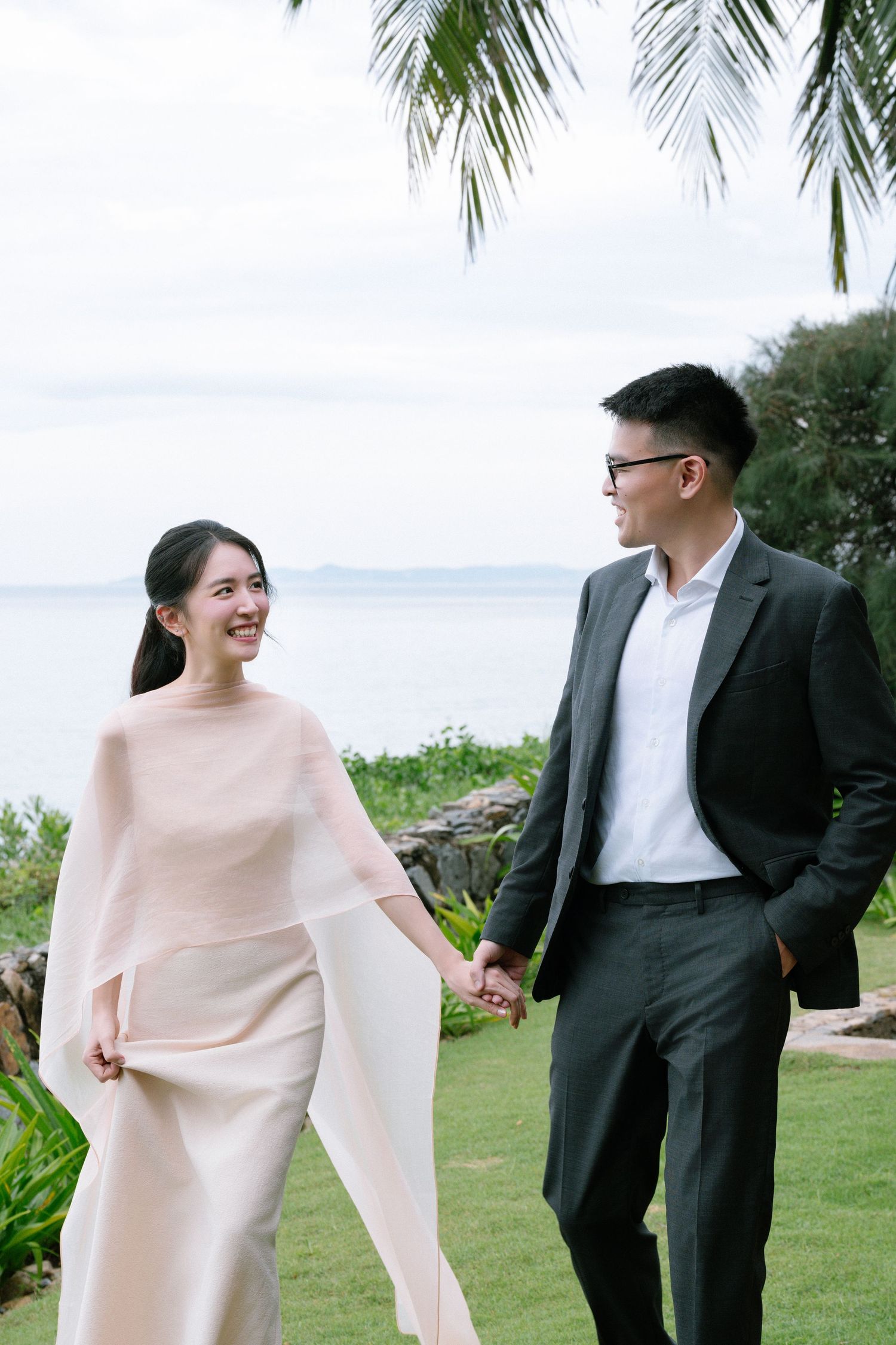 Smiling couple holding hands in garden with ocean backdrop during engagement photoshoot.