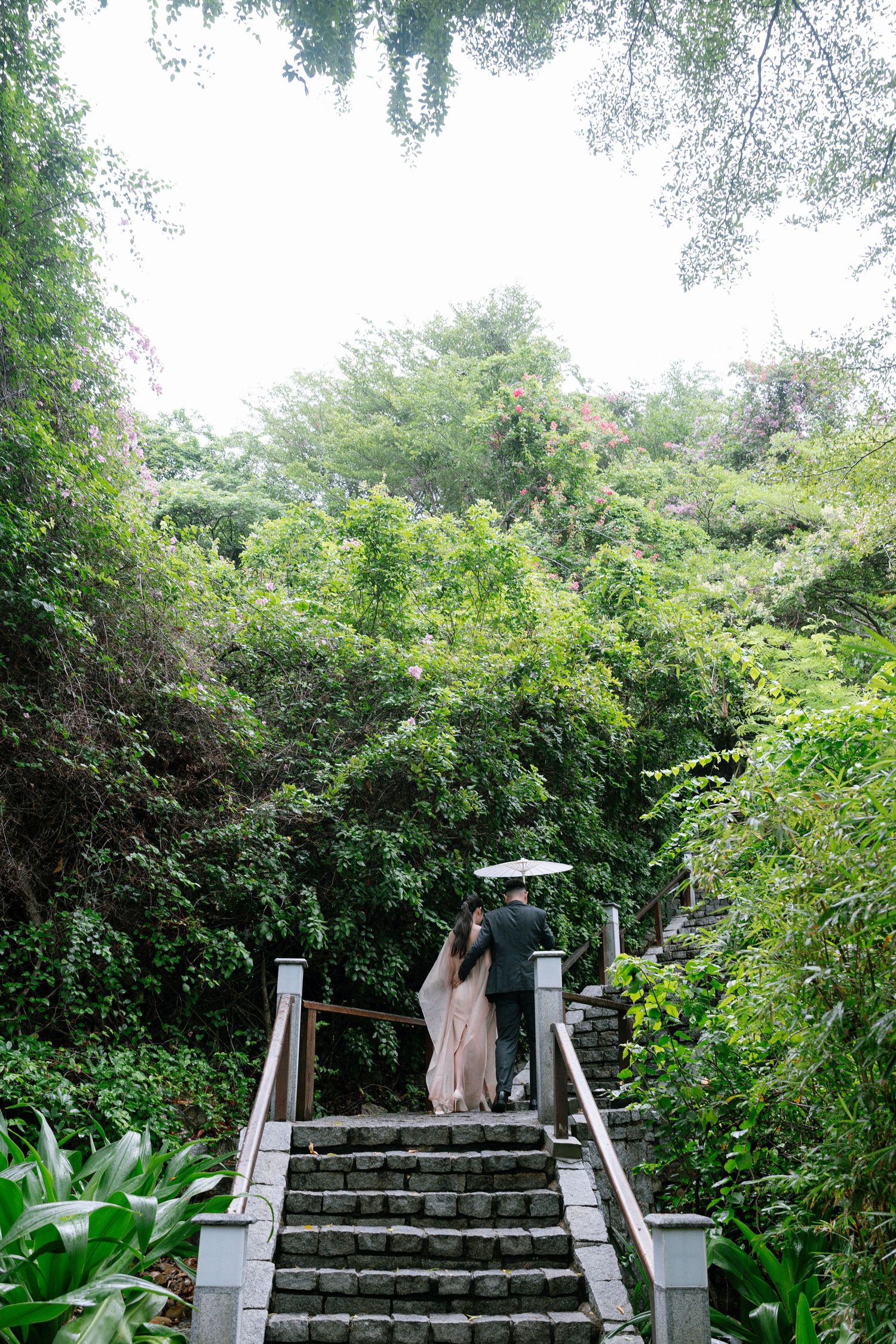 Bride and groom walking up stone steps through lush tropical greenery with umbrella.