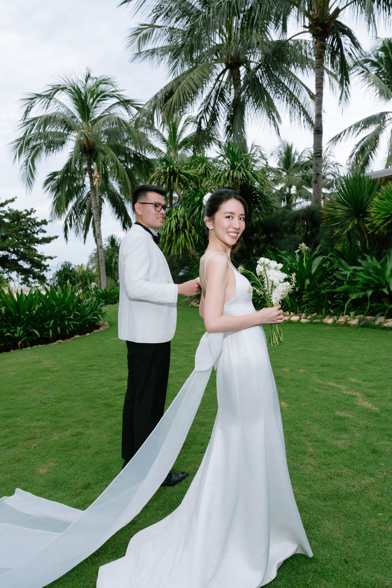 Groom in white tuxedo adjusting bride's elegant white gown on tropical garden lawn.