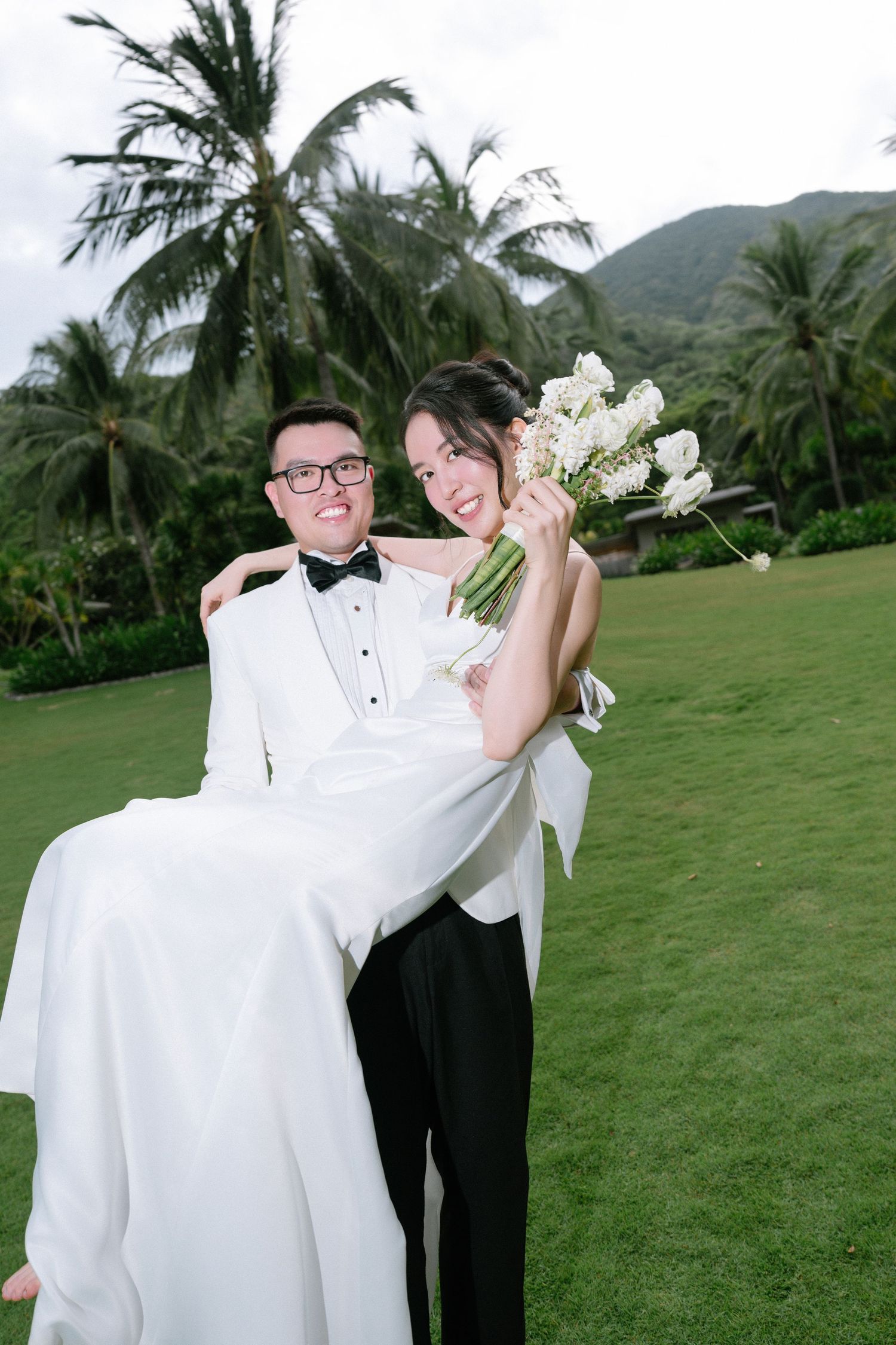 Joyful groom carrying laughing bride holding bouquet on lush green lawn with palms.