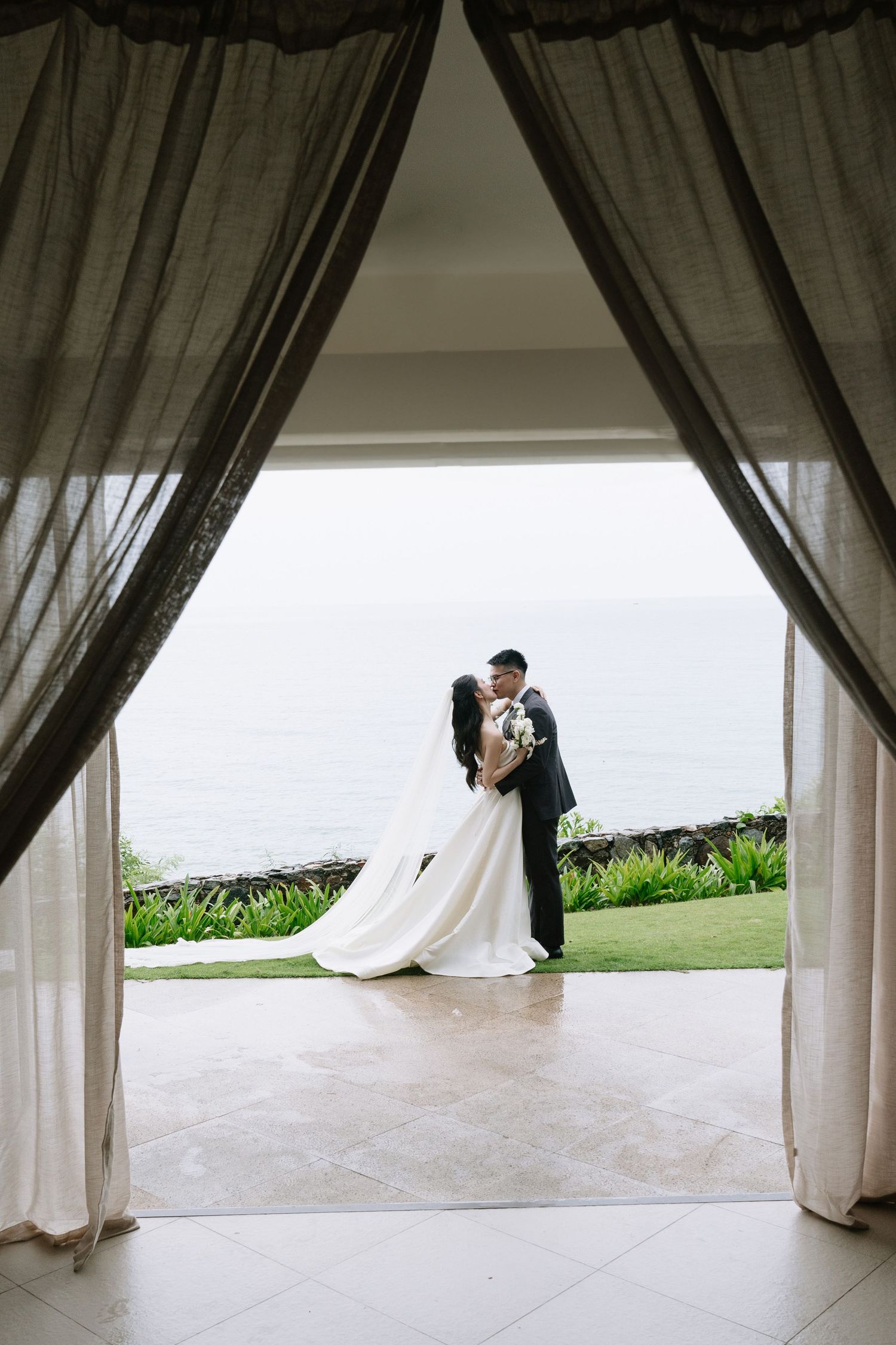 Bride and groom sharing a kiss framed by curtains with ocean view behind them.