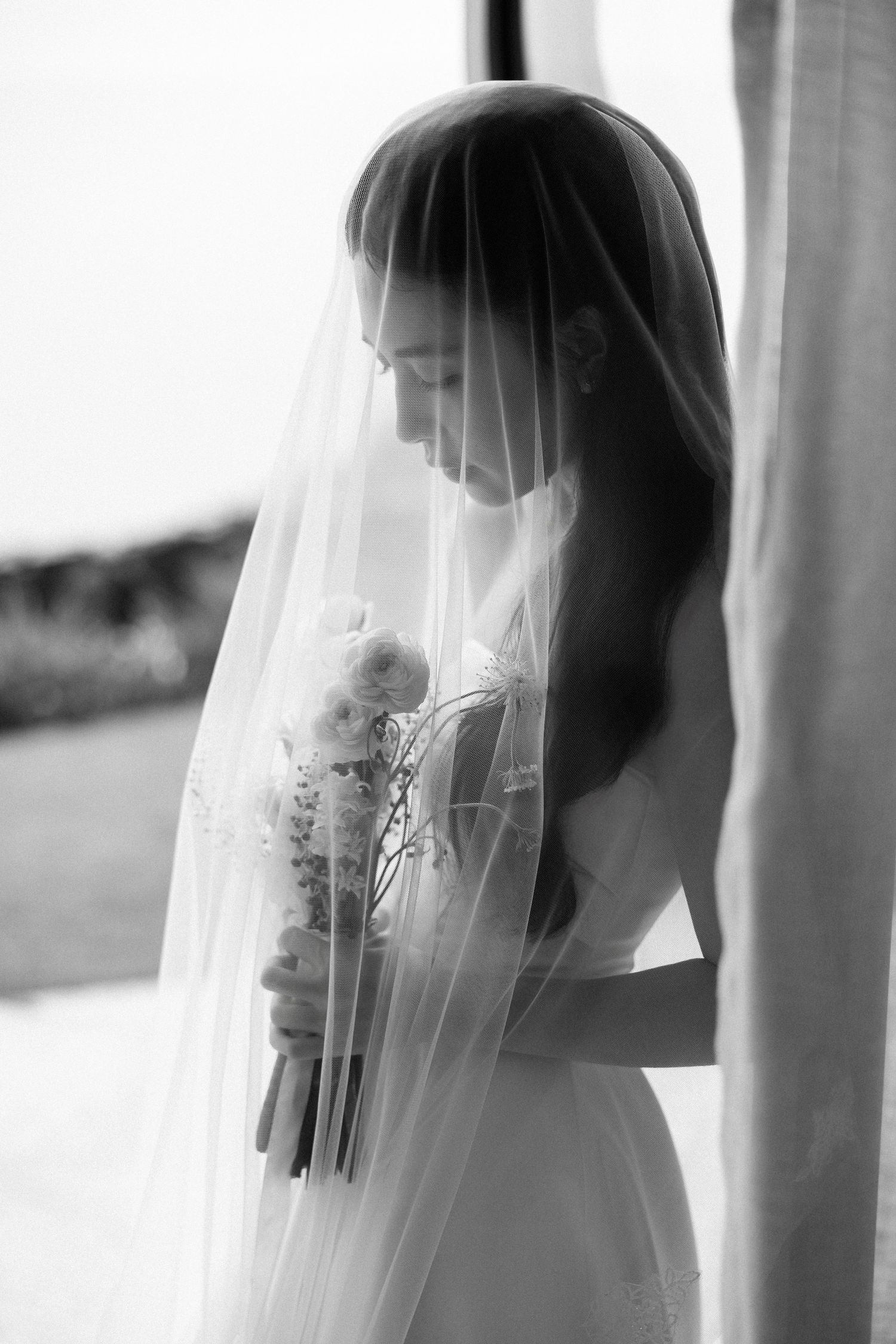 Black and white portrait of veiled bride holding bouquet in soft window light.