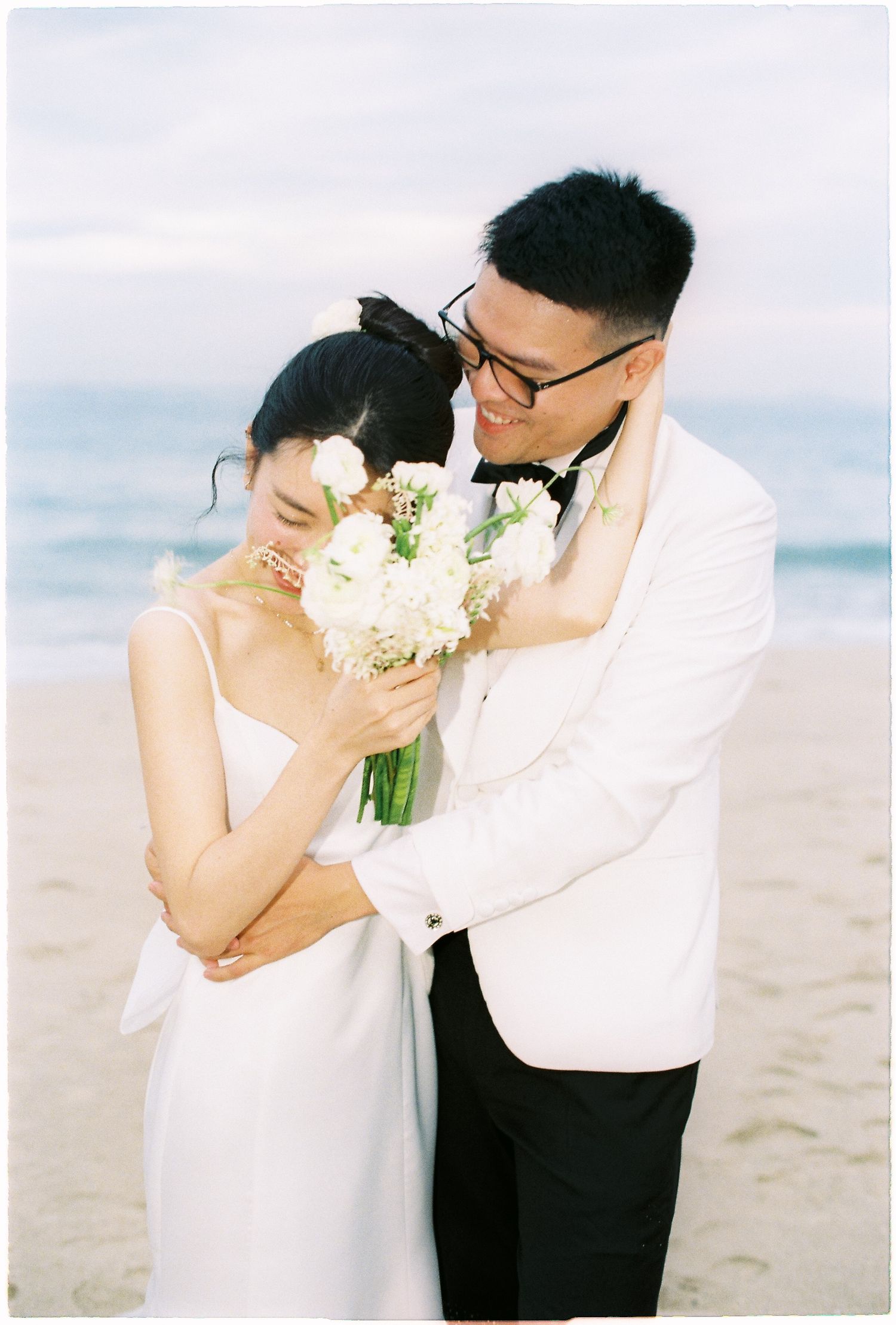 Groom in white tuxedo embracing bride holding white bouquet on romantic beach setting.