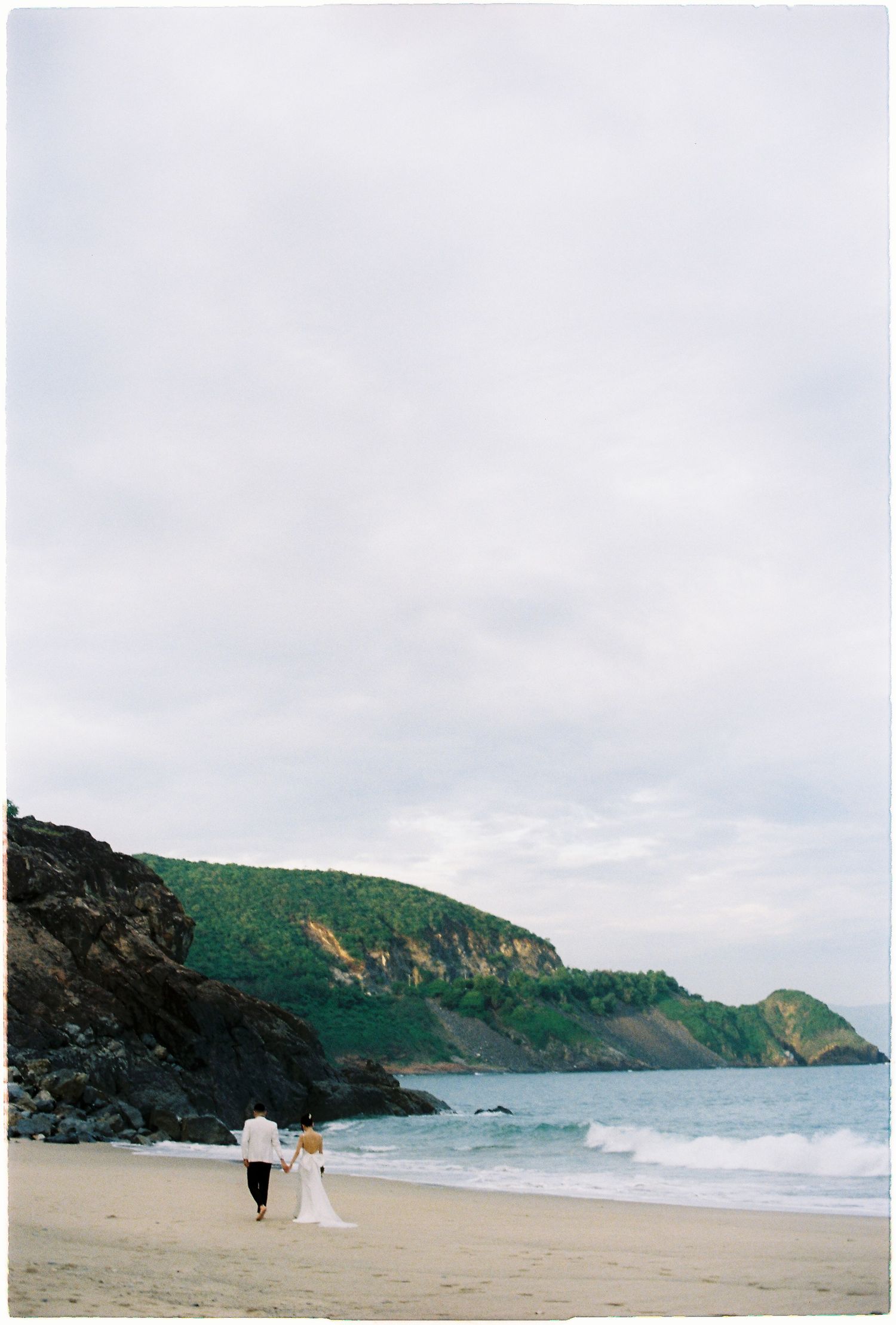 Bride and groom walking hand in hand along a scenic beach with rocky cliffs and ocean waves.
