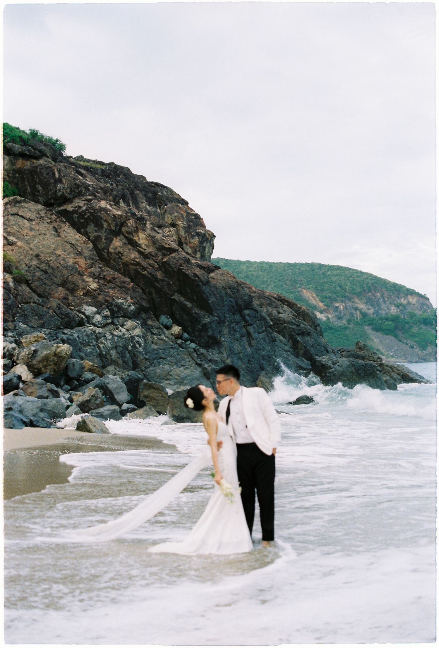 Wedding couple standing in ocean waves beside dramatic rocky cliffs during beach photo session.