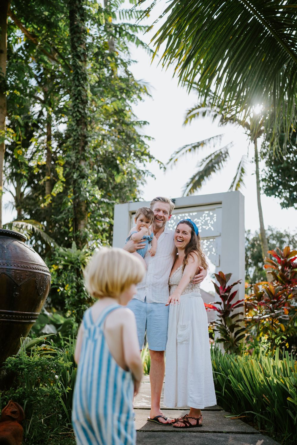 Candid family photo session in a lush tropical garden with white gates and palm trees.