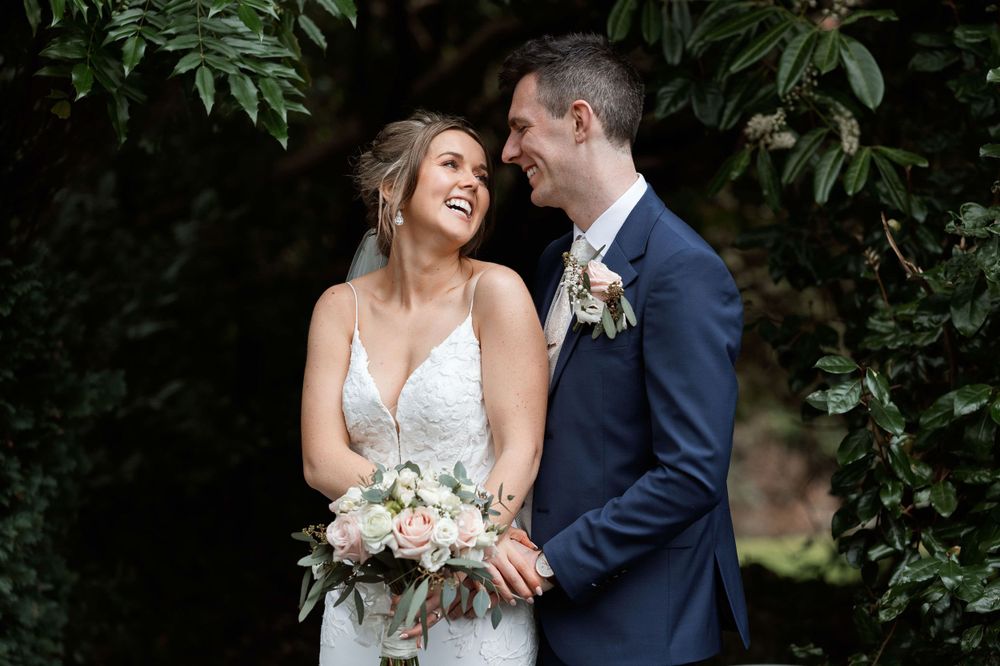Romantic wedding moment captured outdoors with elegant white dress and navy suit against lush greenery backdrop.