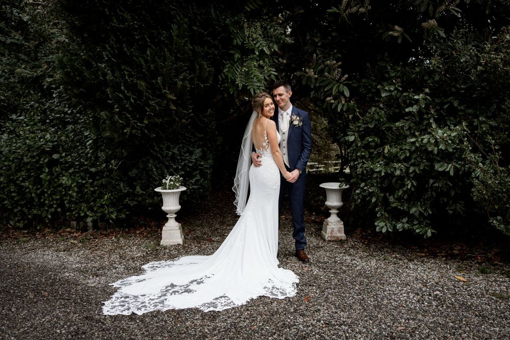 Wedding couple poses between stone pillars with bride's long lace train trailing on gravel path.