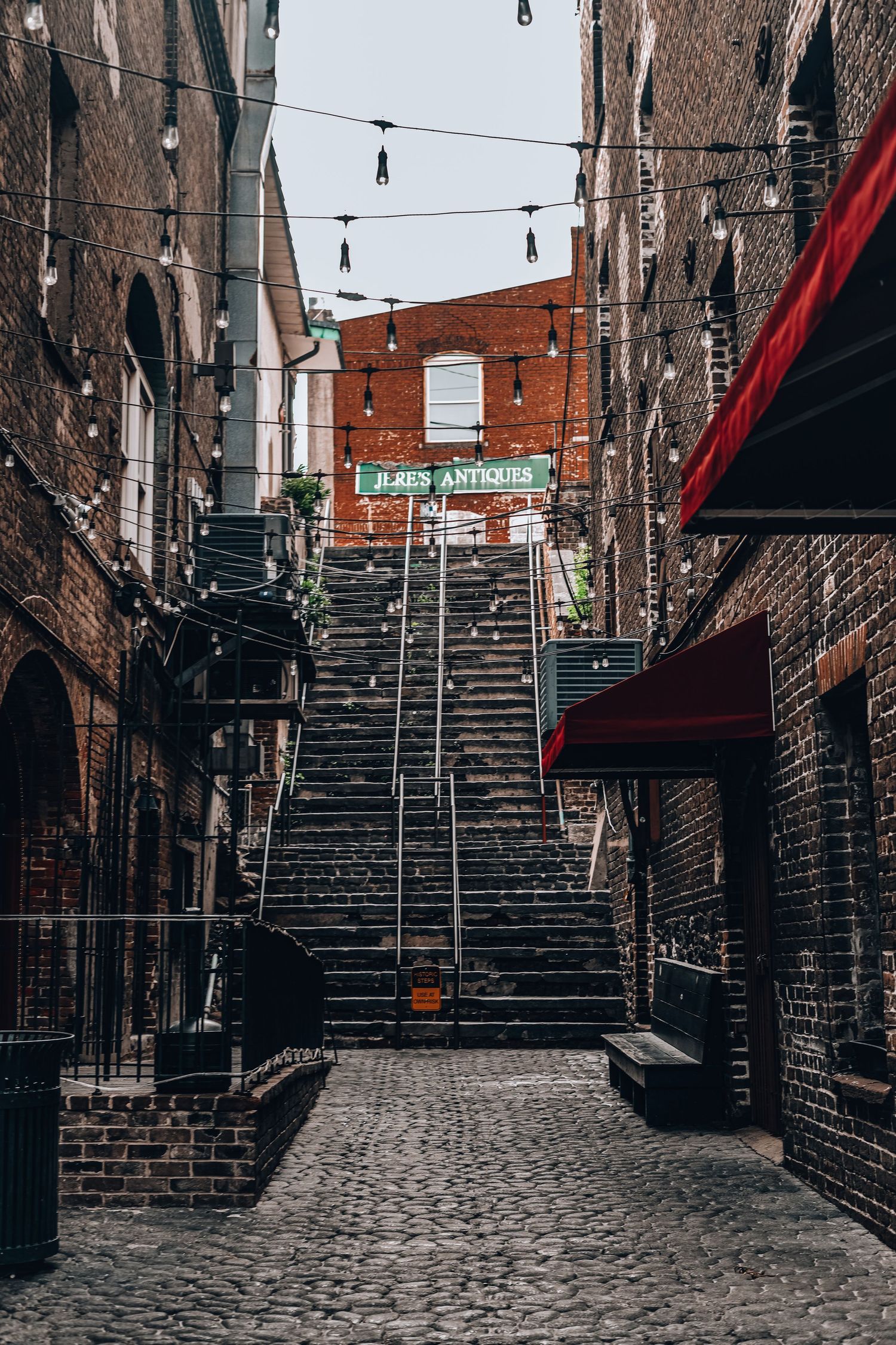 Steep brick staircase between historic buildings with hanging lights and red awnings.