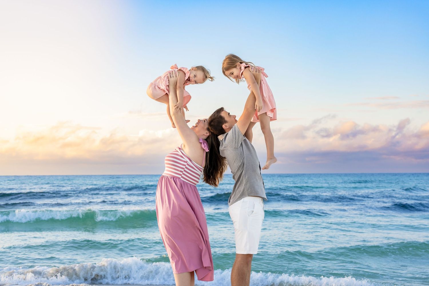 Family in coordinated pink and white outfits playing together on a sandy beach at sunset.