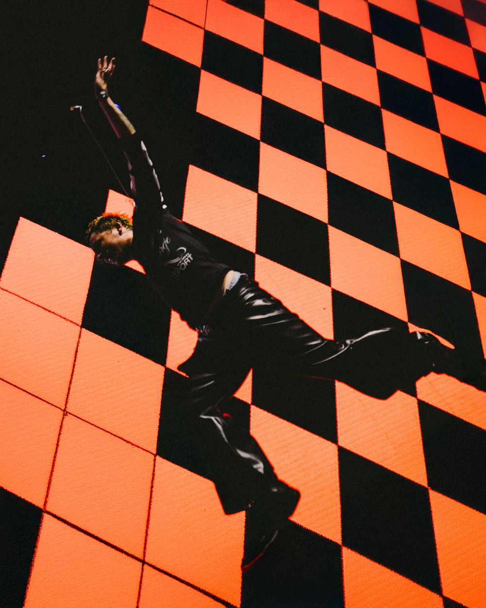 A person in black clothing strikes a dramatic pose against an orange and black checkered wall in dramatic lighting.
