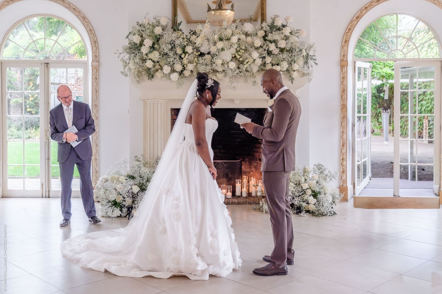 Elegant wedding ceremony in a bright white room with floral arch and fireplace as two people exchange vows.