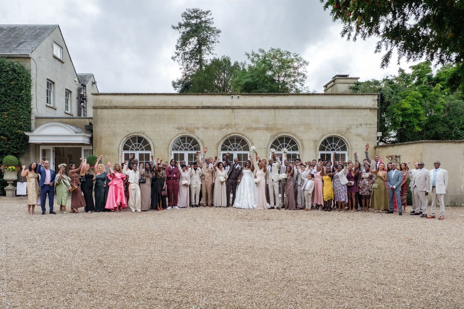 Wedding guests lined up for a group photo outside a historic building on a sunny day.