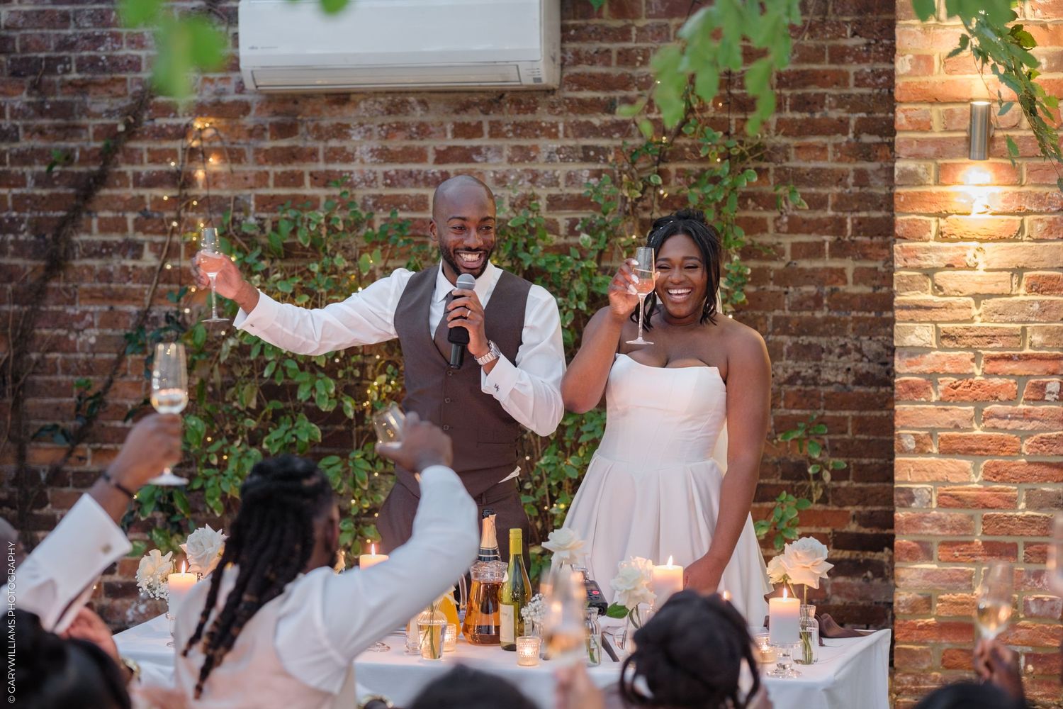 Wedding guests celebrate and dance together at an indoor reception with exposed brick walls and string lights.