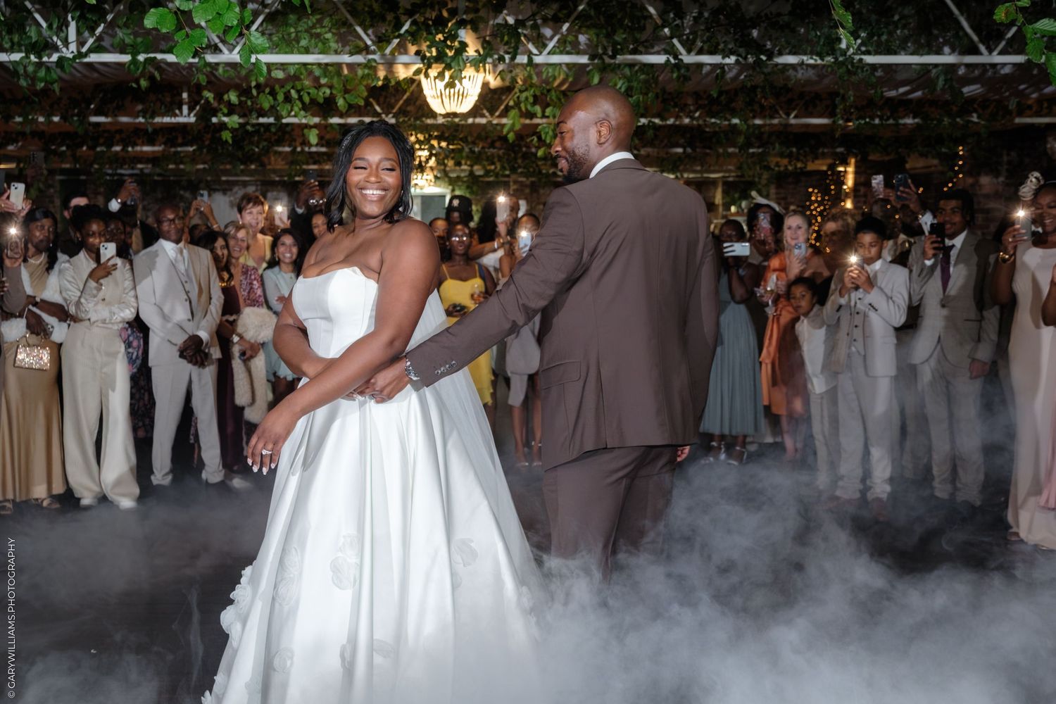 Wedding guests dance together in a reception hall decorated with string lights and greenery.