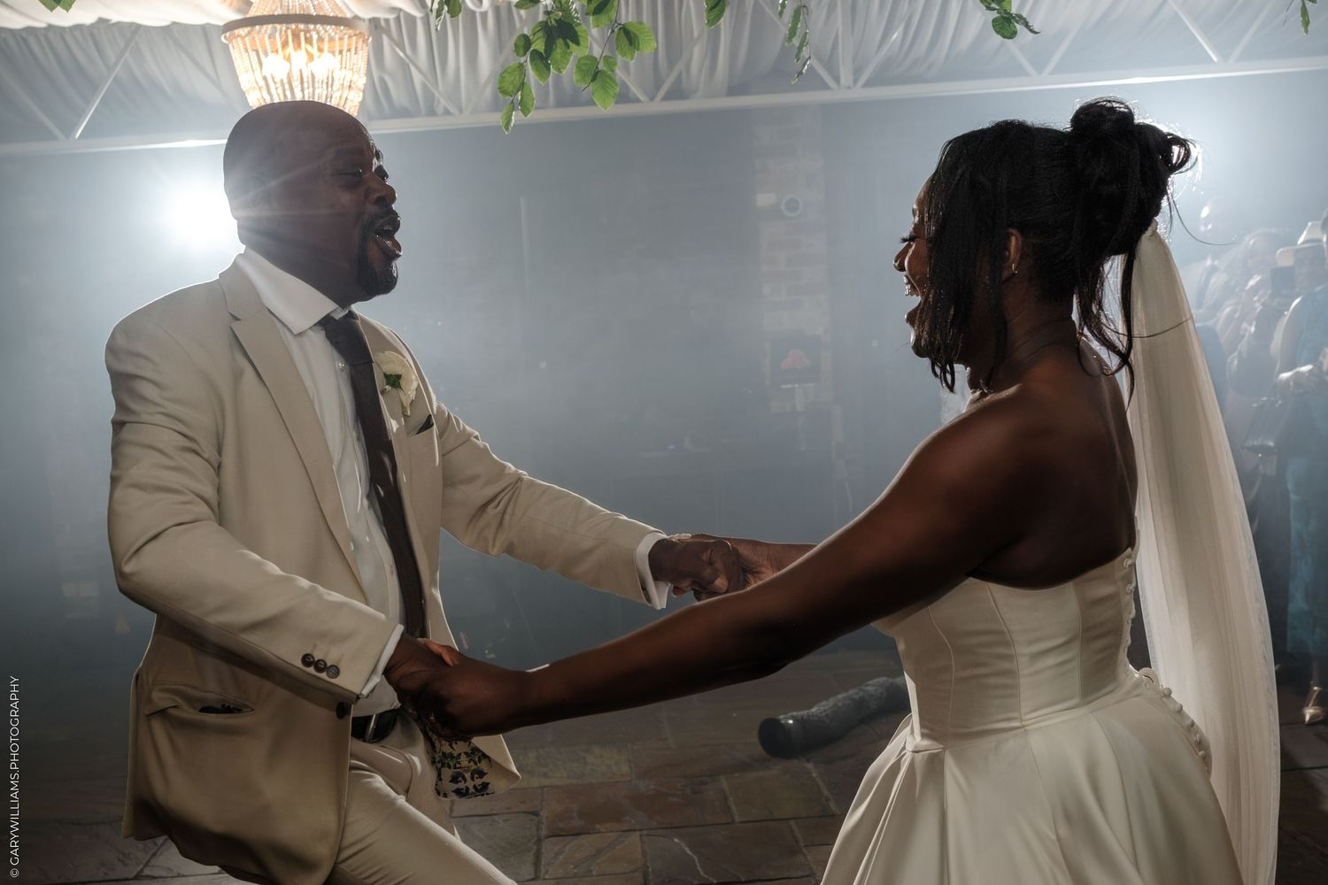 A couple in formal wedding attire embraces during their first dance at a reception with soft lighting and smoke effects.