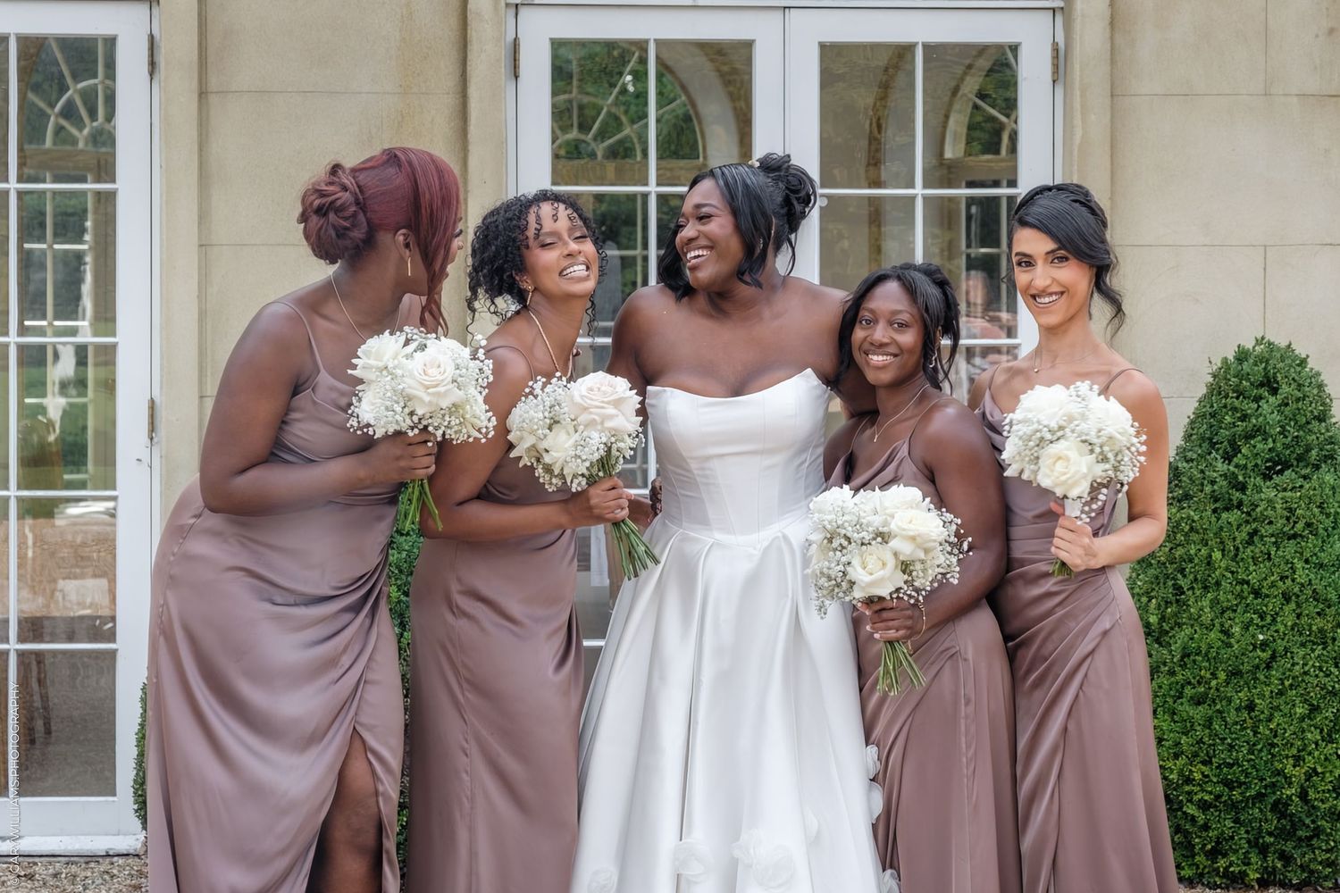 Bridesmaids in dusty rose dresses share a joyful moment with bride in white gown holding white bouquets.