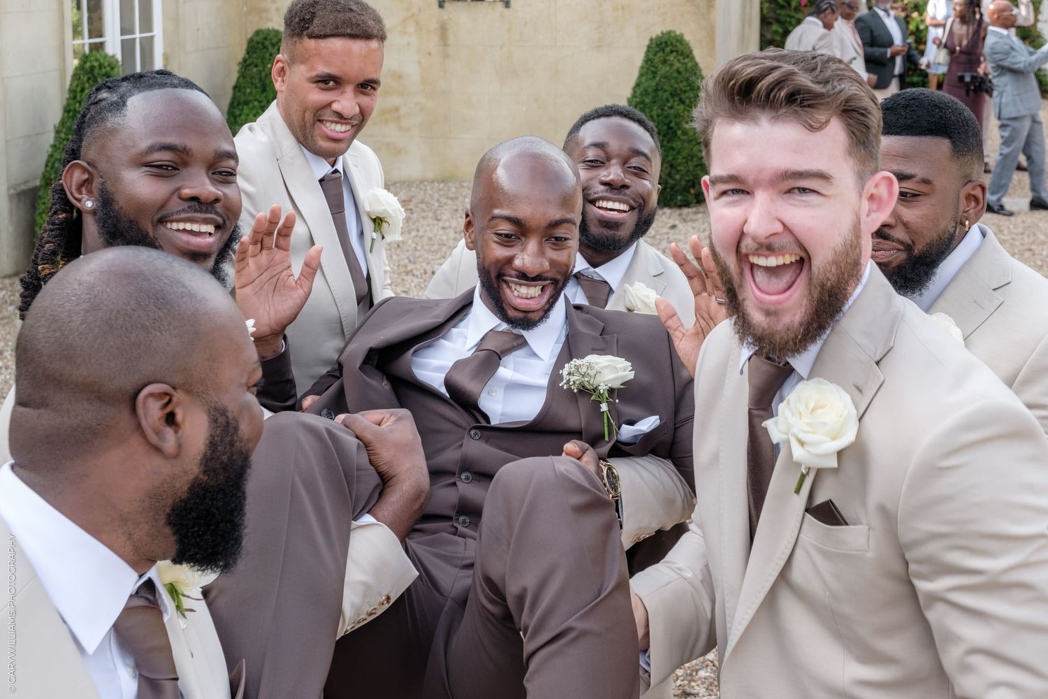 Wedding party members in suits laugh and celebrate together during outdoor photo session.