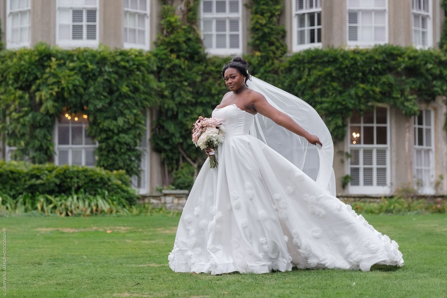 Person in flowing white wedding dress walking across lush green lawn outside stone building with ivy-covered walls.