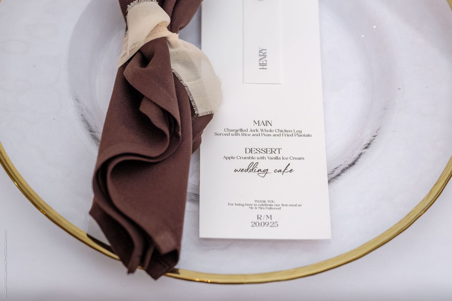 Elegant place setting with brown napkin and menu card on white plate with gold rim detail.