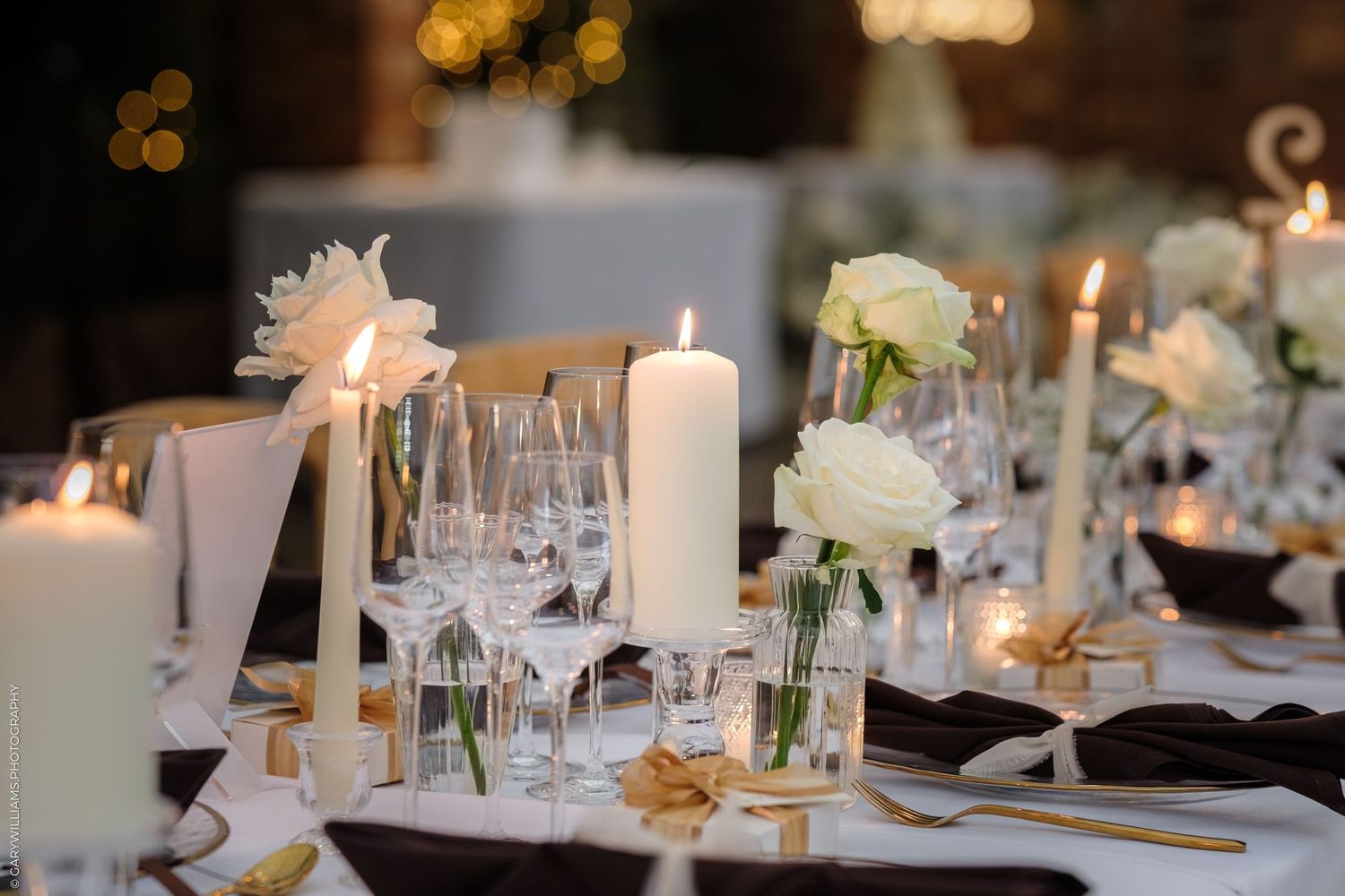 Elegant wedding reception tables decorated with white candles, wine glasses and floral centerpieces in soft lighting.
