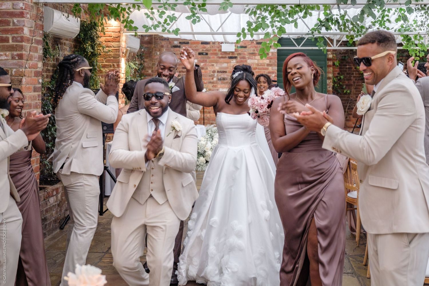 Wedding party in neutral beige and brown attire dances together in celebration at outdoor brick venue.