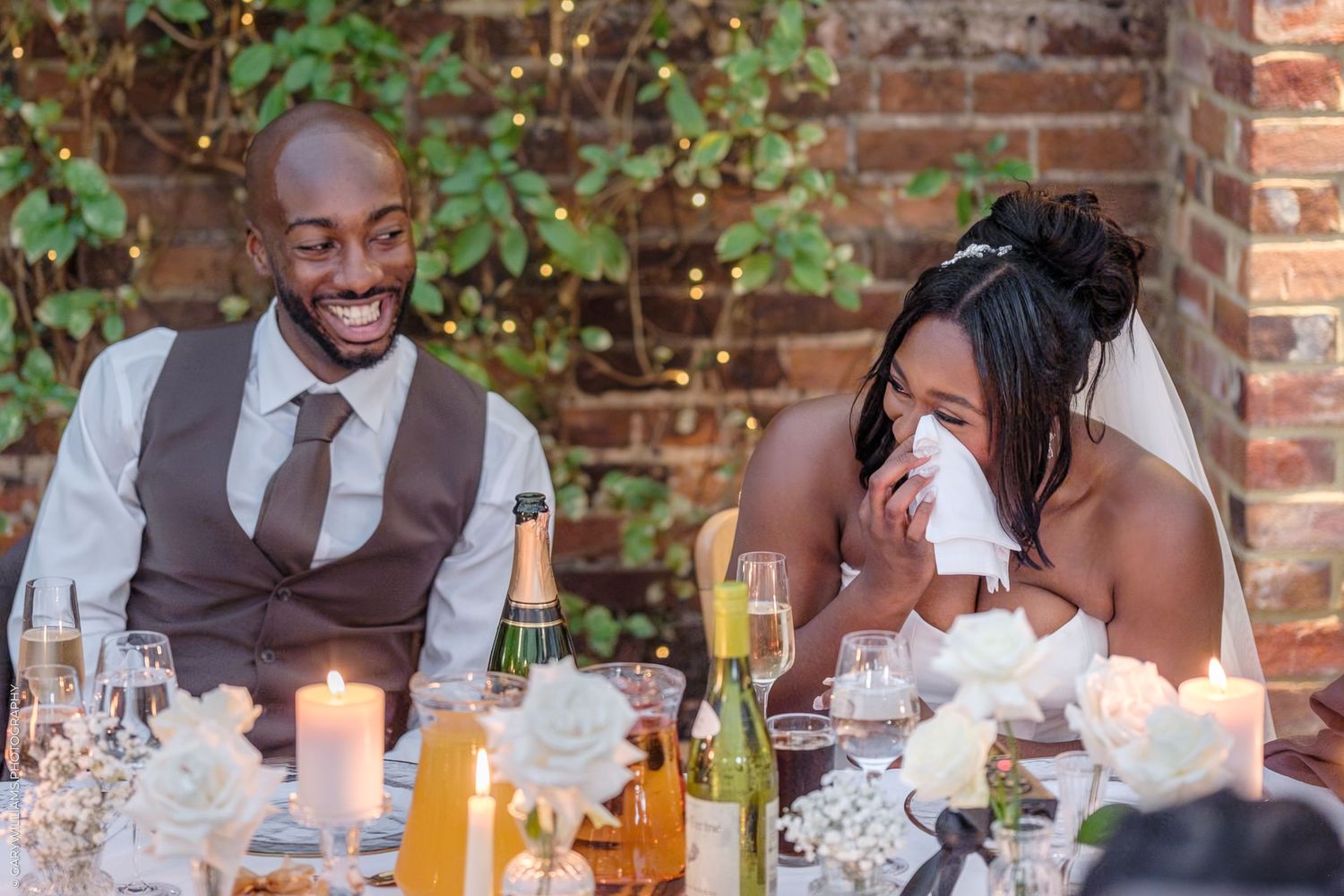 Wedding guests share joyful laughter during an outdoor reception at a rustic brick venue.