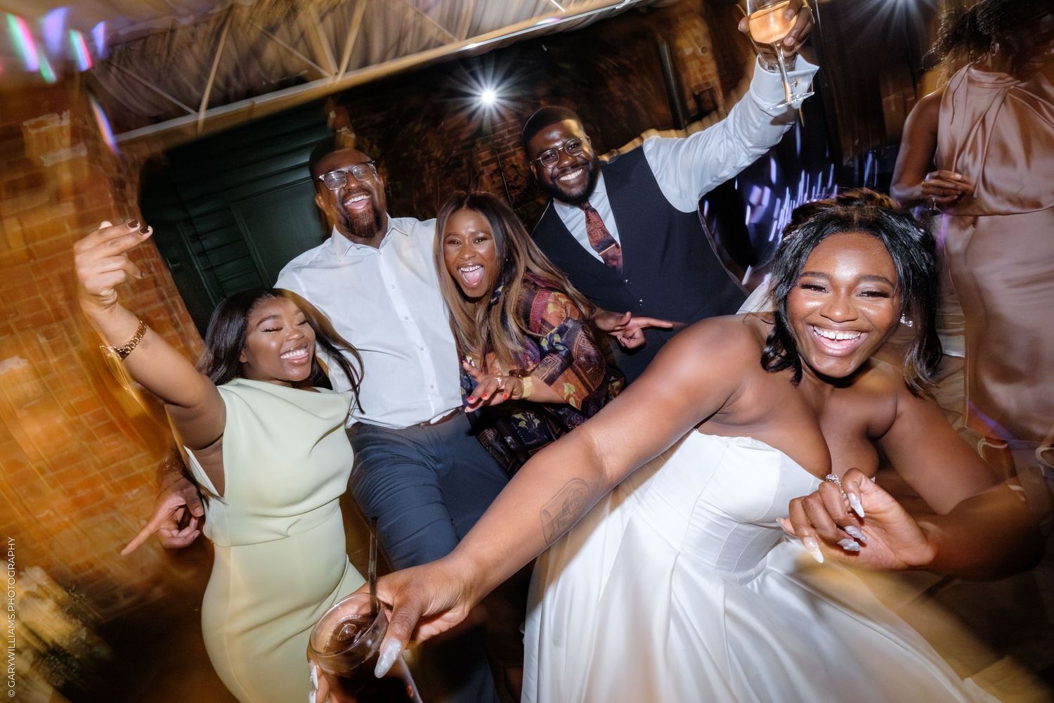 Wedding guests dance joyfully together on a wooden dance floor at an evening reception celebration.