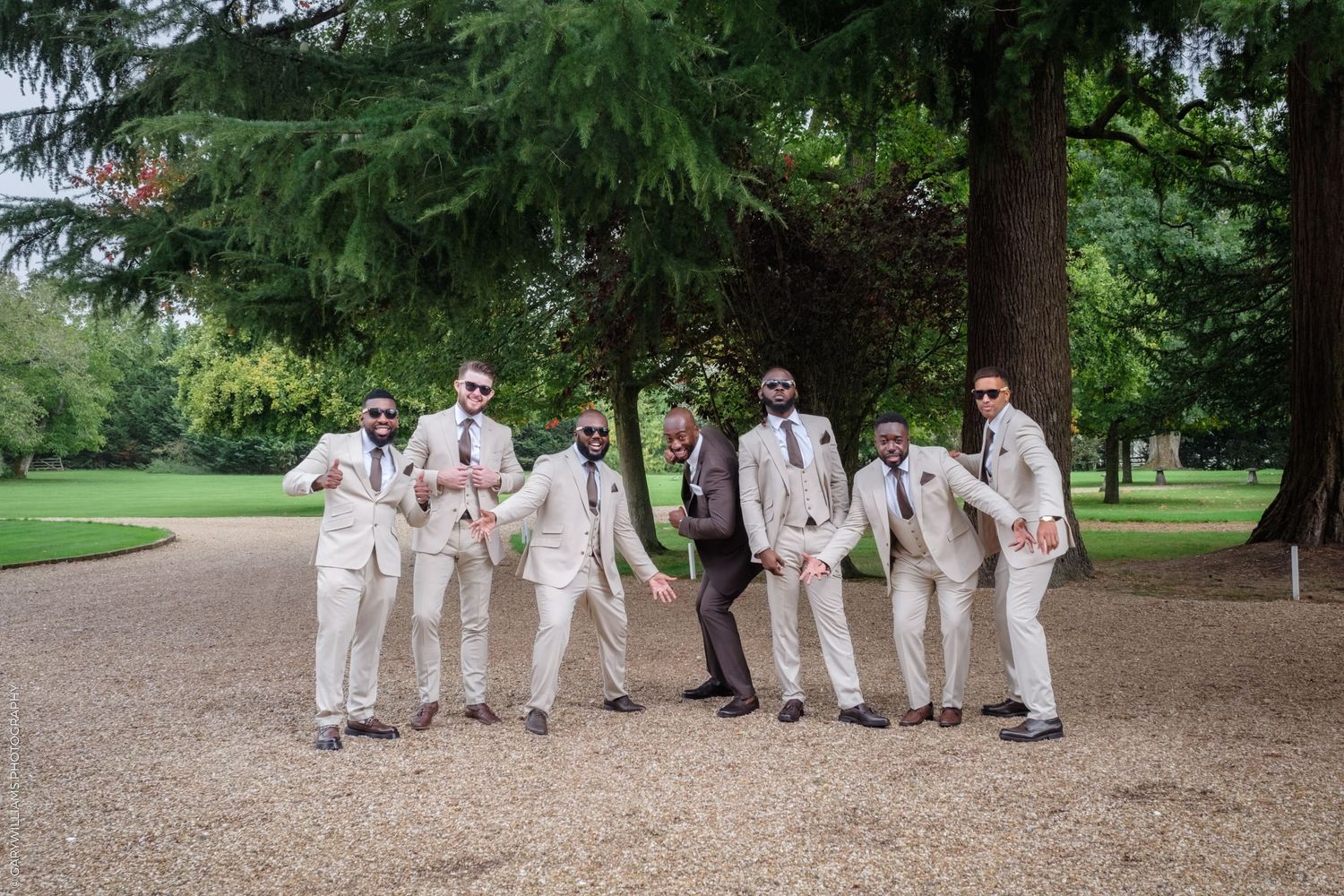 Wedding party in white attire poses together under tall trees in a scenic outdoor garden setting.