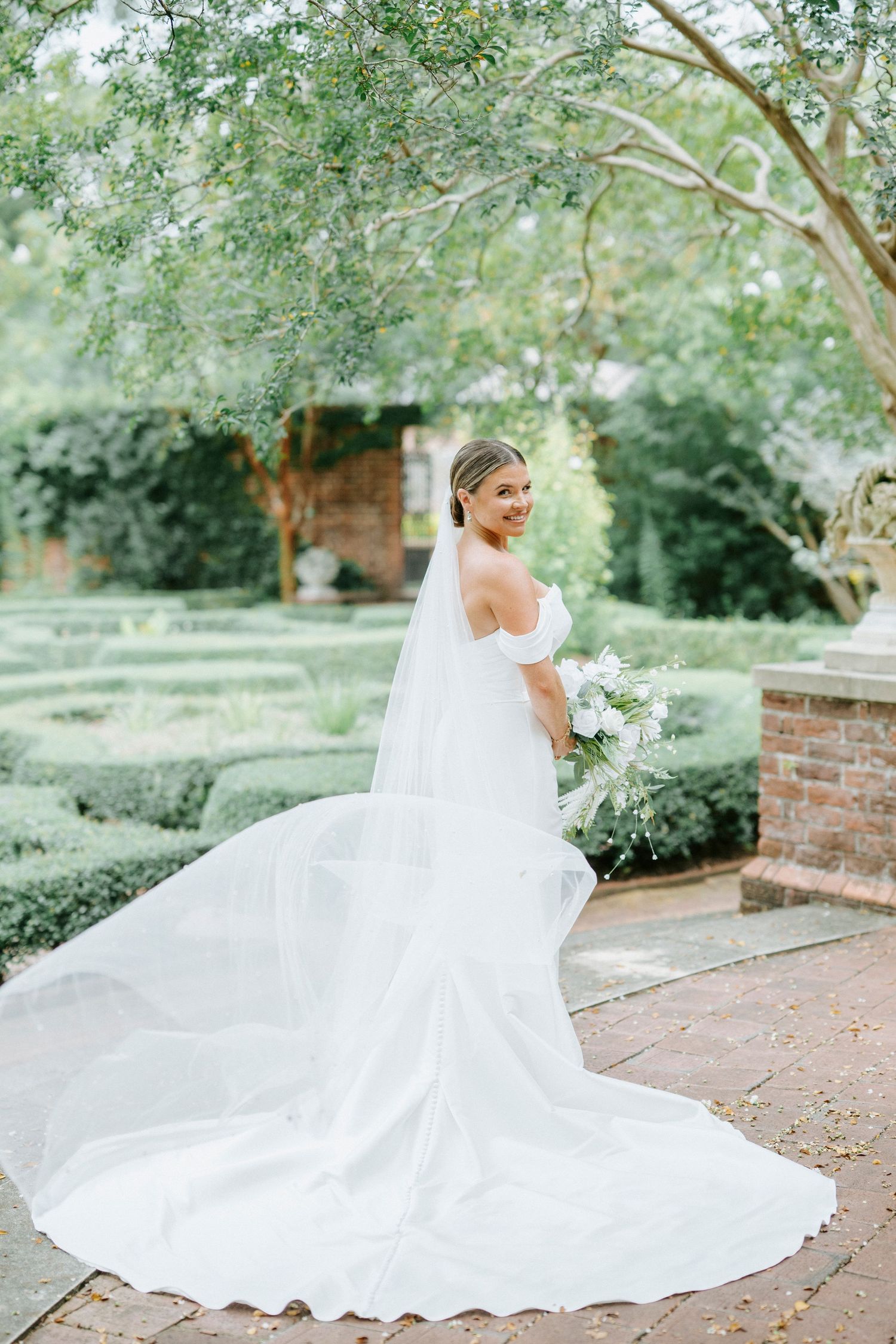 Beautiful bridal portrait in a garden setting with flowing white dress and long veil against lush greenery and brick walls.