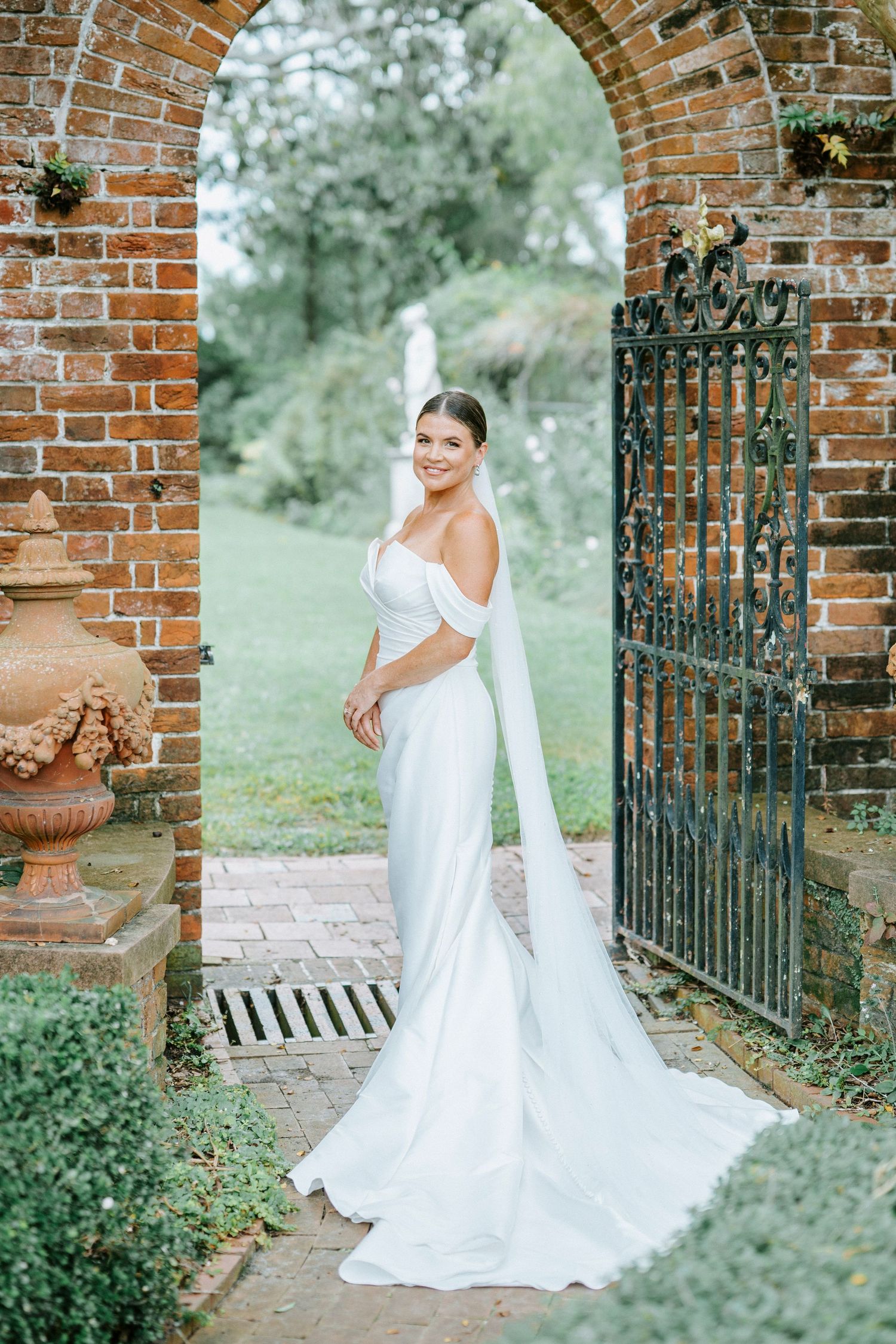 A bride in a white wedding gown poses gracefully under a rustic brick archway surrounded by greenery.