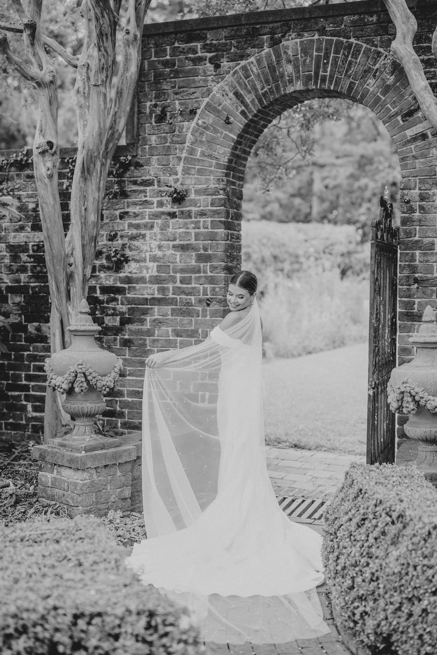 A flowing white wedding veil blows in the wind against a rustic brick wall and garden backdrop.