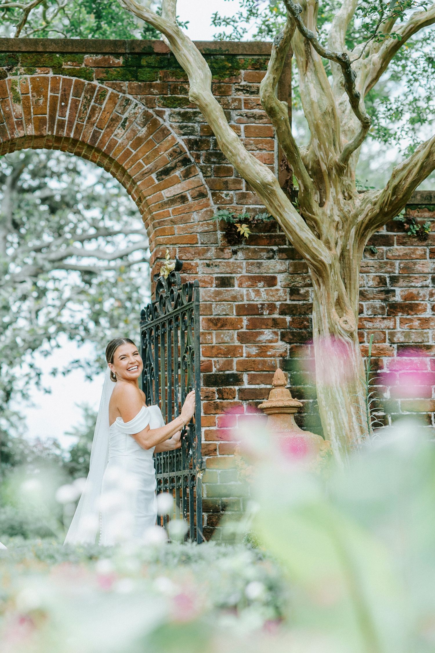 A figure in a white dress poses elegantly against a rustic brick archway surrounded by greenery and pink flowers.